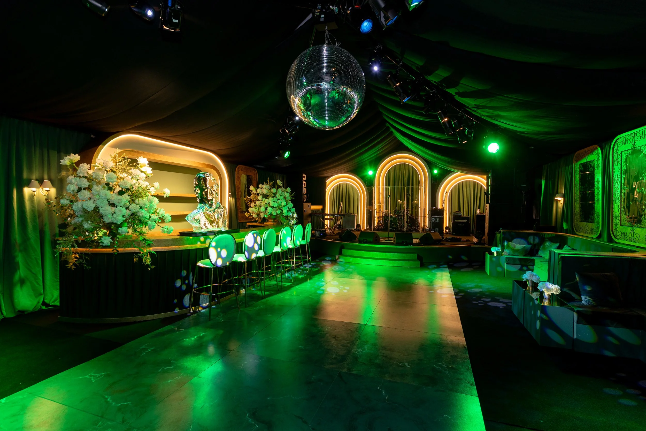 An empty event space with a stage, floral arrangements, and seating area, decorated with green lighting and a disco ball hanging from the ceiling.