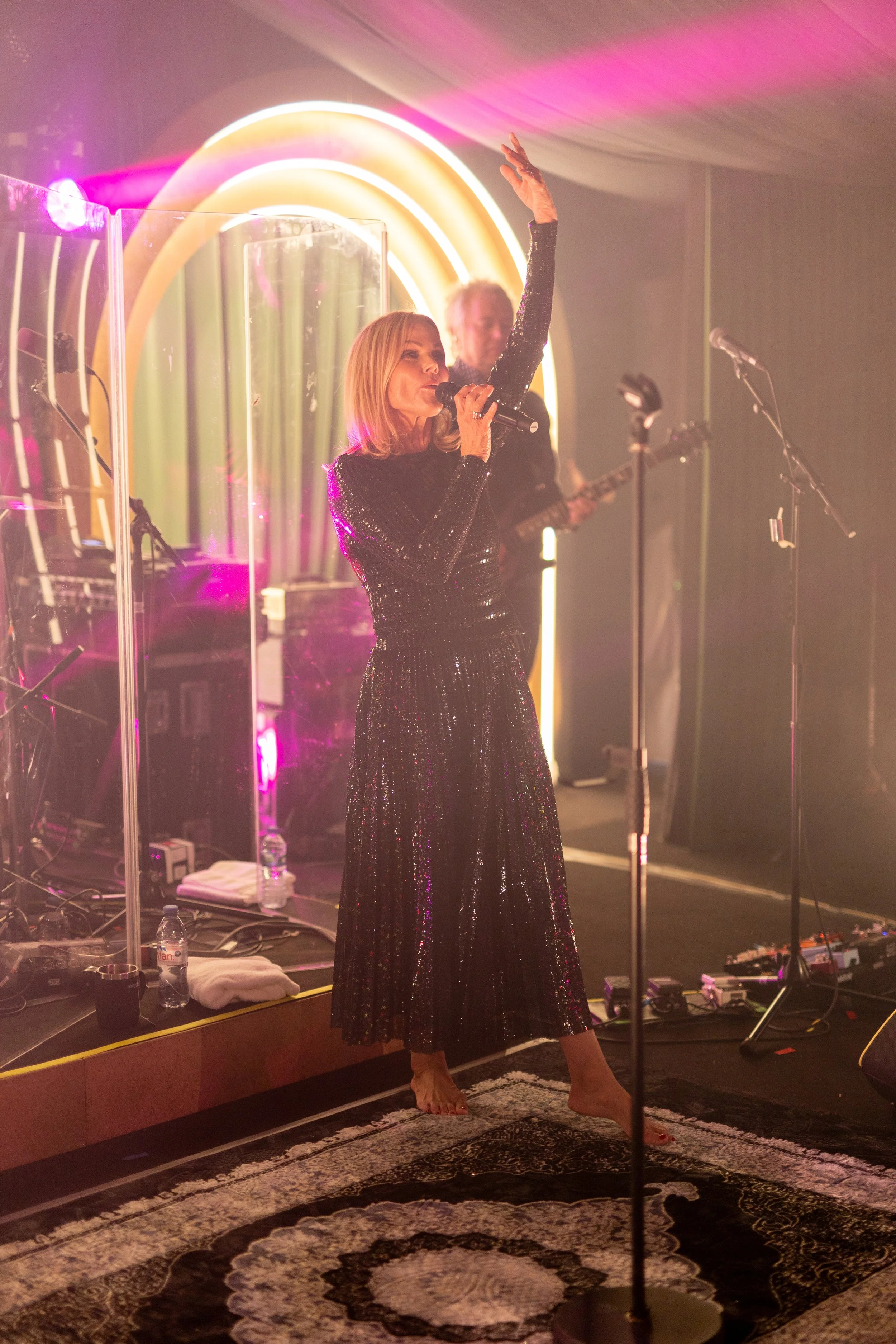A woman in a black, sparkly dress sings into a microphone on stage, with her arm raised, during a performance. A musician plays guitar behind her, with neon rainbow lights in the background.