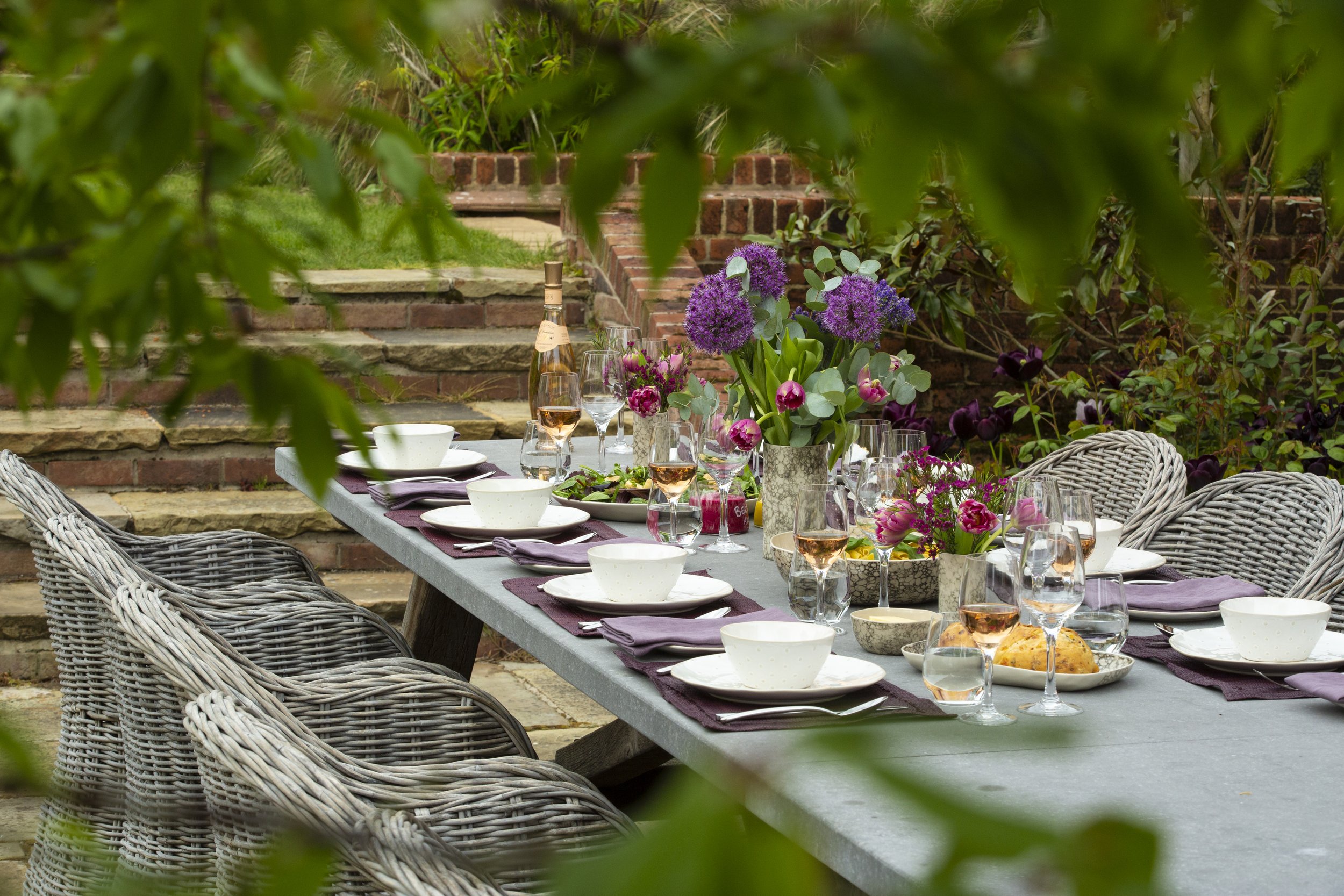 A long outdoor dining table set with plates, glasses, and purple napkins, decorated with vases of purple and pink flowers, situated on a stone patio with brick steps and greenery in the background.