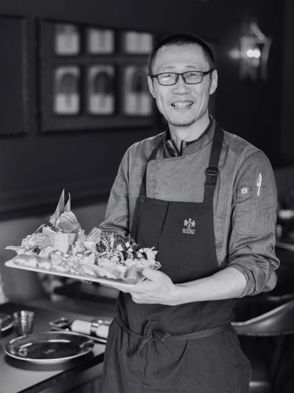 A smiling man in glasses and a chef's apron holding a tray of decorated desserts in a restaurant kitchen.