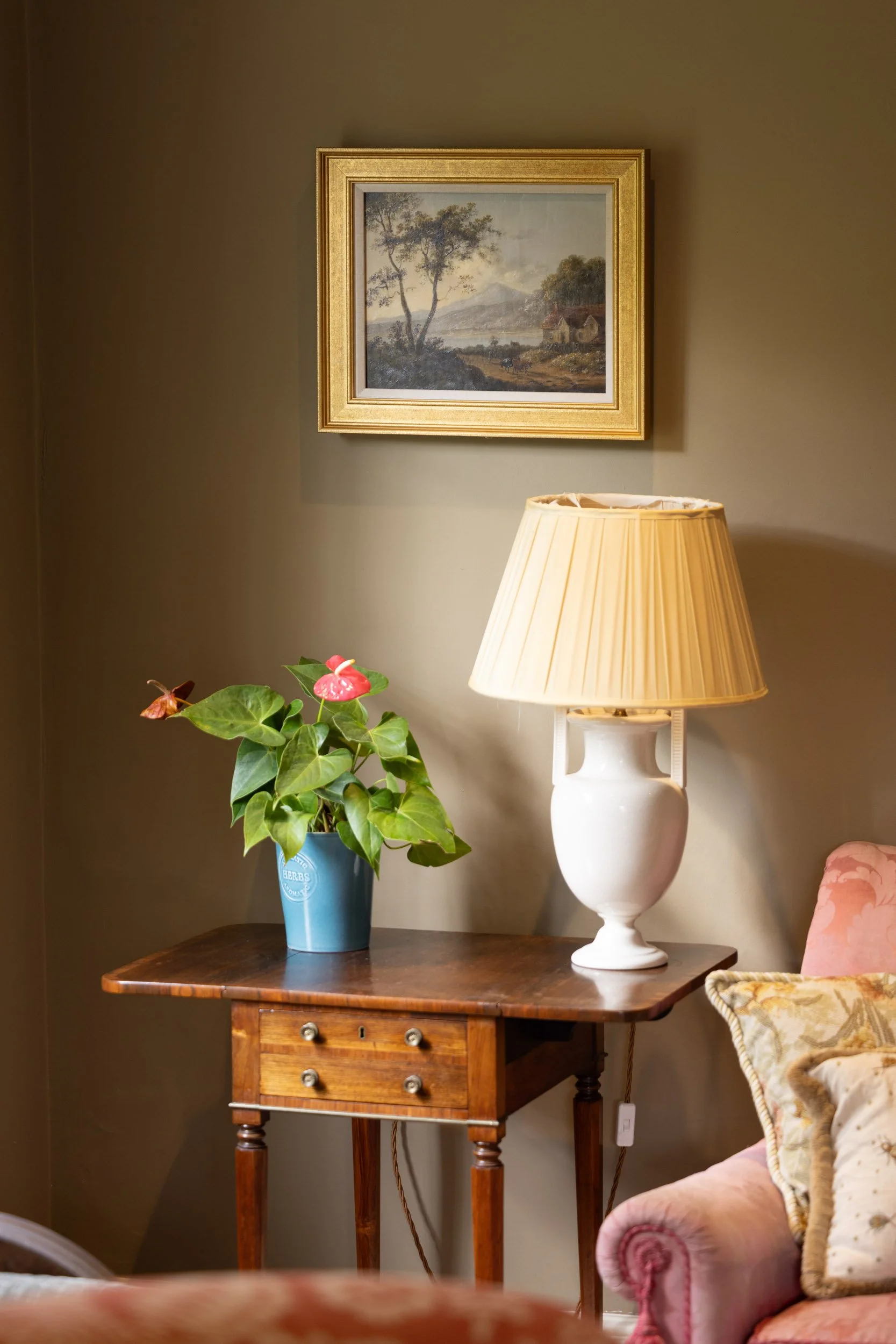 A wooden side table with small drawers and brass hardware, holding a blue herb pot and a white ceramic lamp with a beige lampshade, against a beige wall with framed landscape painting.
