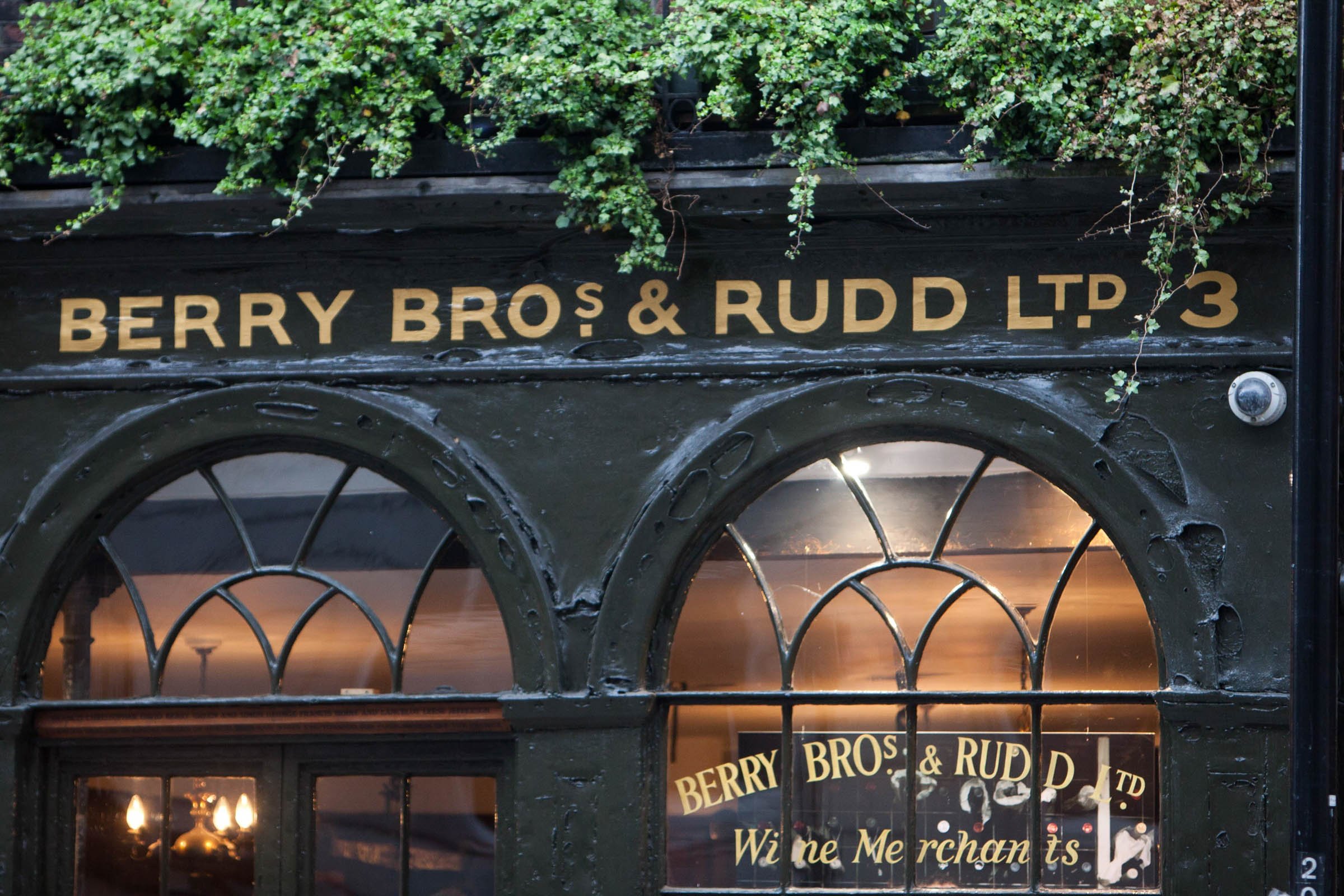 Sign with the text 'BERRY BROS & RUDD LTD' above a window, and a reflection of the same text in the glass. Green foliage with small leaves above the sign and window.