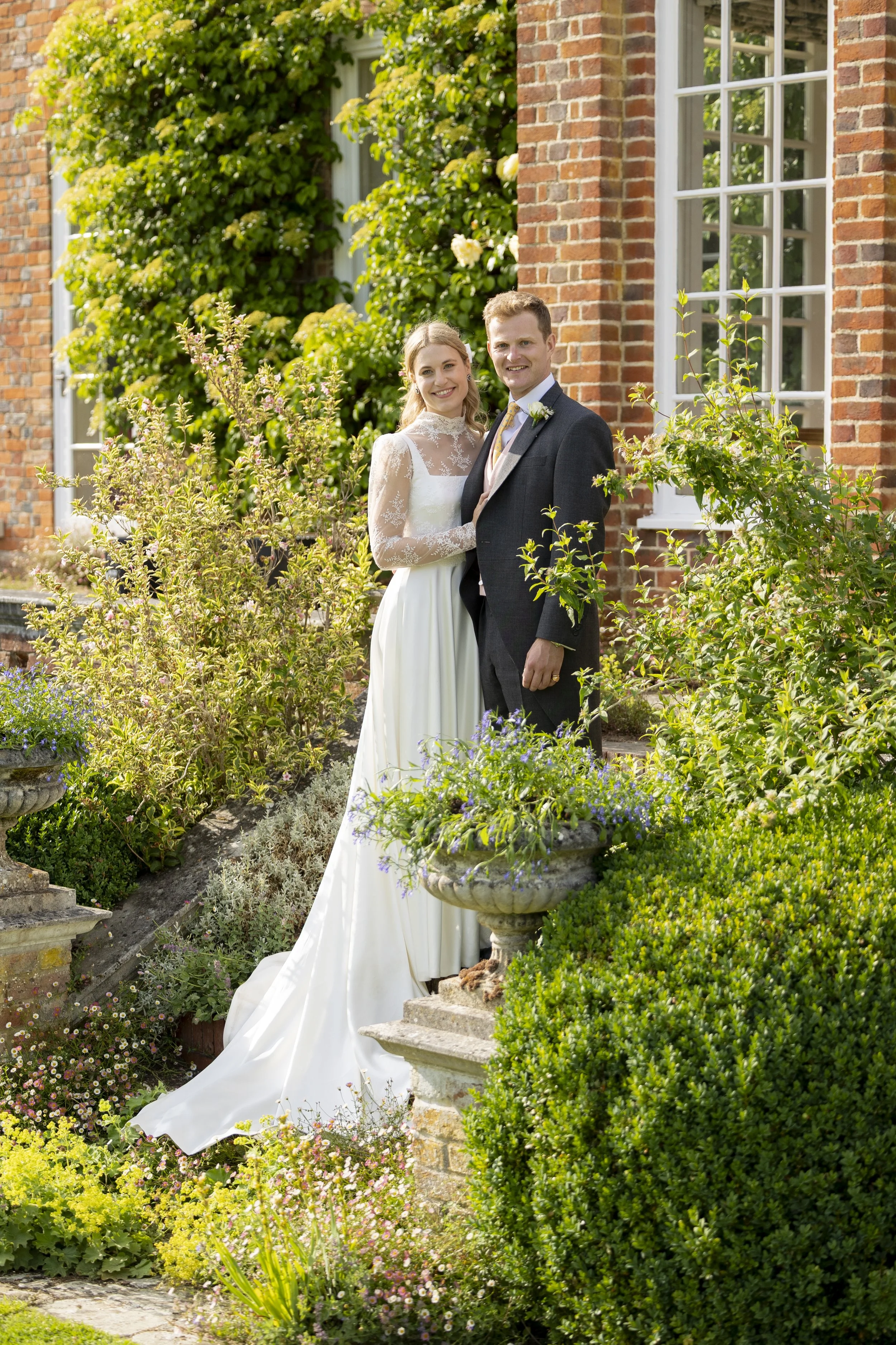 A newlywed couple in wedding attire posing outdoors in a lush garden with green bushes, flowering plants, and a brick building with large windows in the background.
