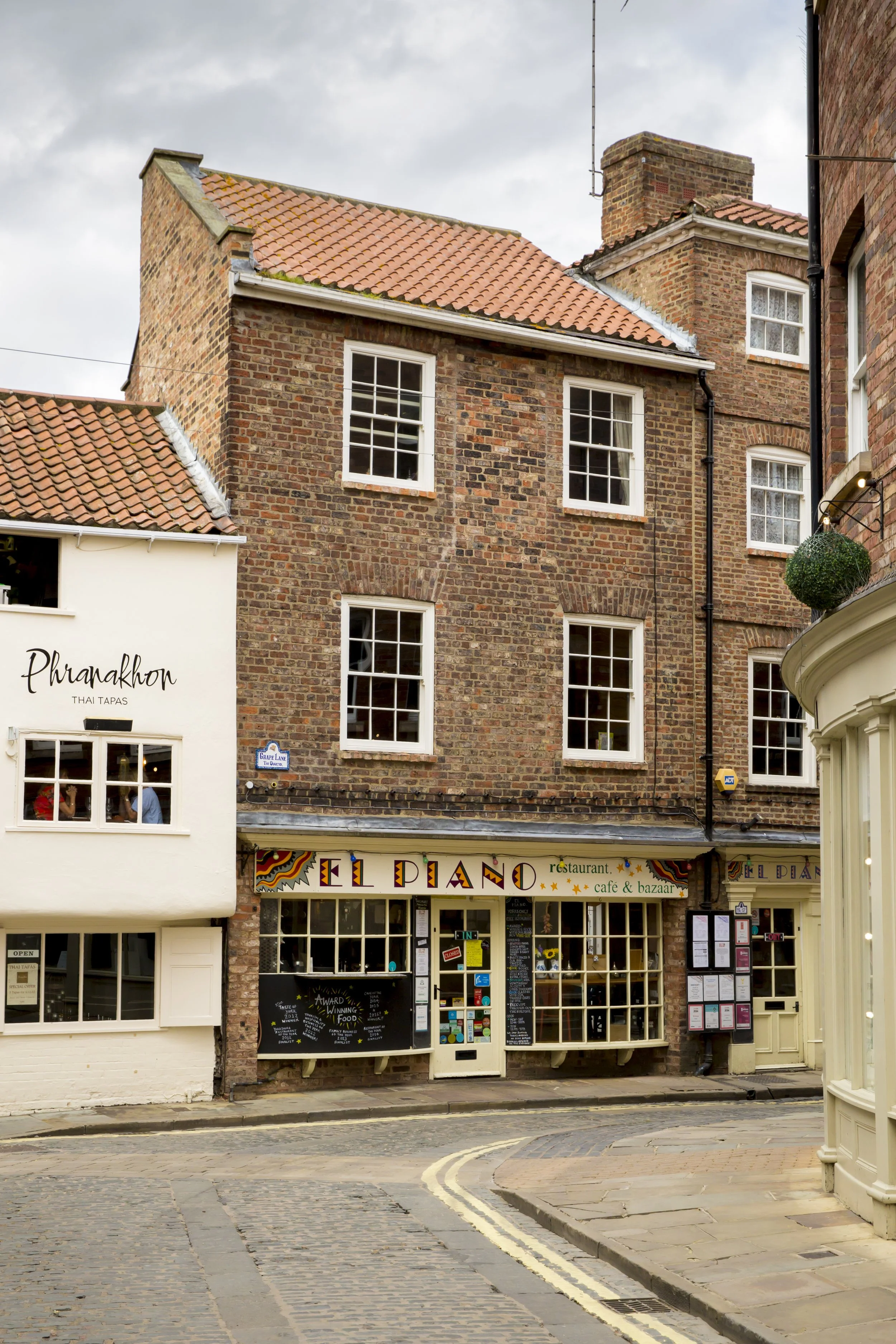 A street view showing a brick building with several windows and a restaurant called 'El Piano' on the ground level, with another building to the left named 'Phranakhon Thai Tapas.'