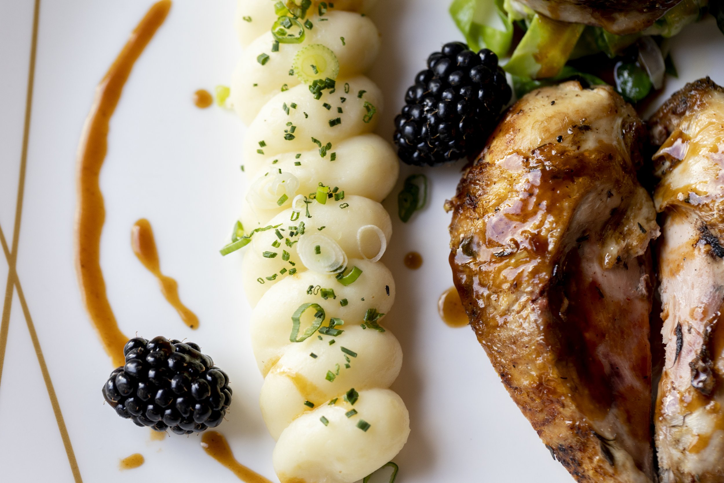 Close-up of a plated dish featuring mashed potatoes garnished with chopped green herbs and sliced green onions, blackberries, and a piece of roasted chicken, with sauce drizzled on the plate.