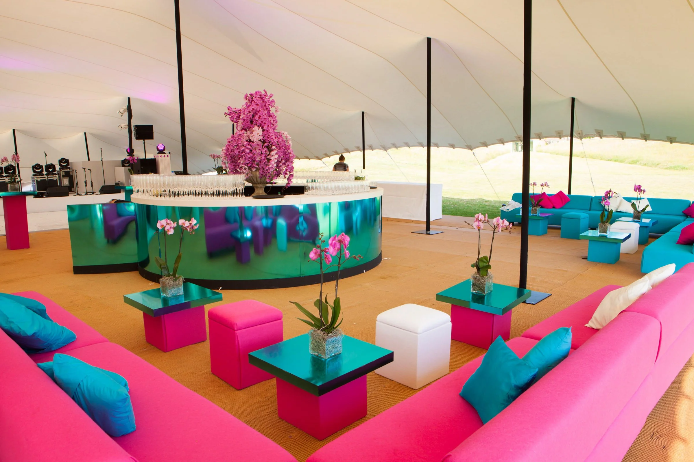 Colorful lounge area with pink and teal furniture, including sofas, pillows, tables, and potted orchids, set under a large white canopy. A bar with pink and purple flowers is in the background.