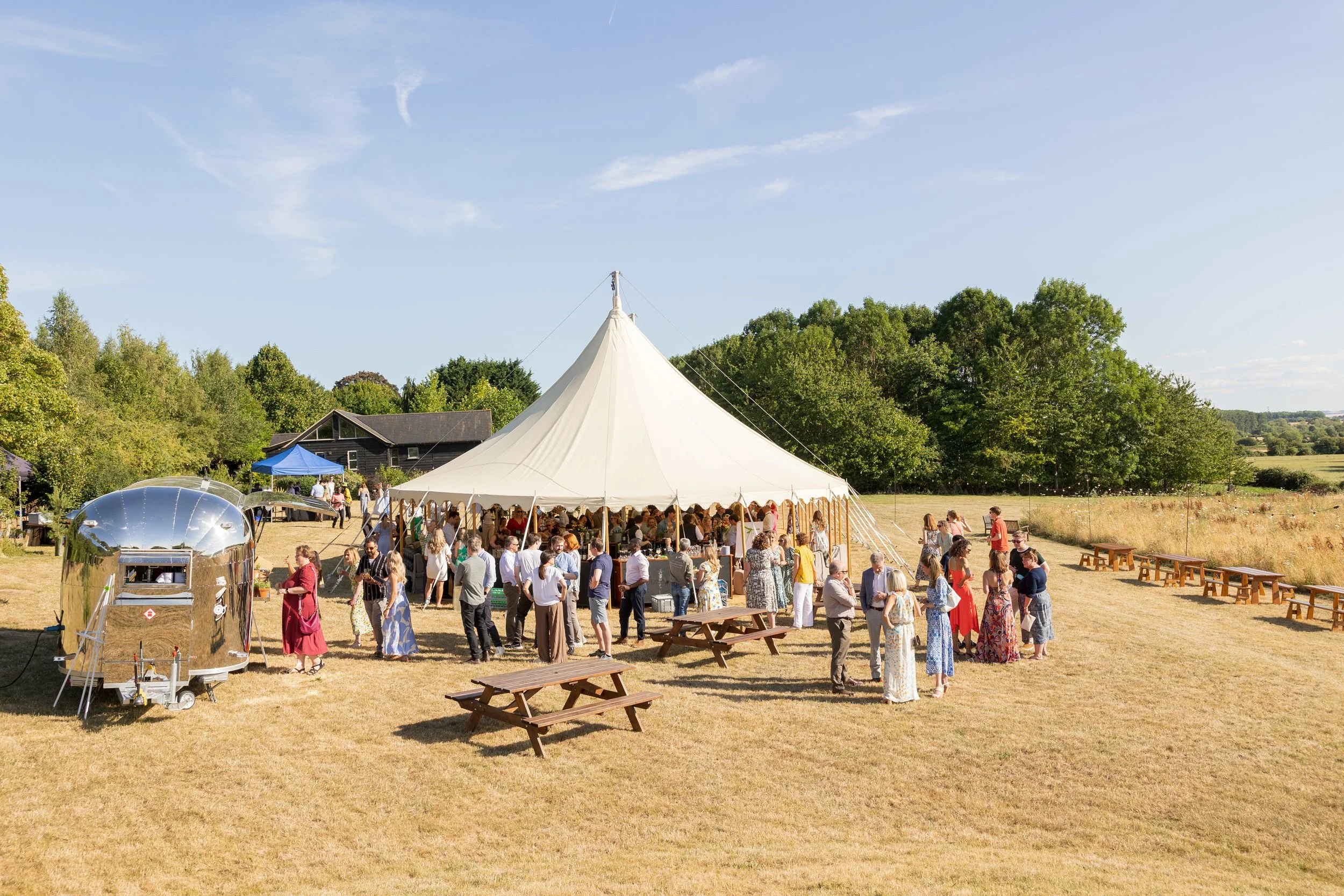 People gathered under a large white tent at an outdoor event on a sunny day, with picnic tables and trees in the background.