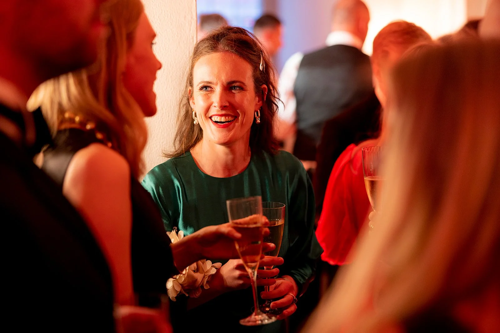 A woman with long brown hair and earrings is smiling and holding a glass of champagne while talking to women at a party or celebration.