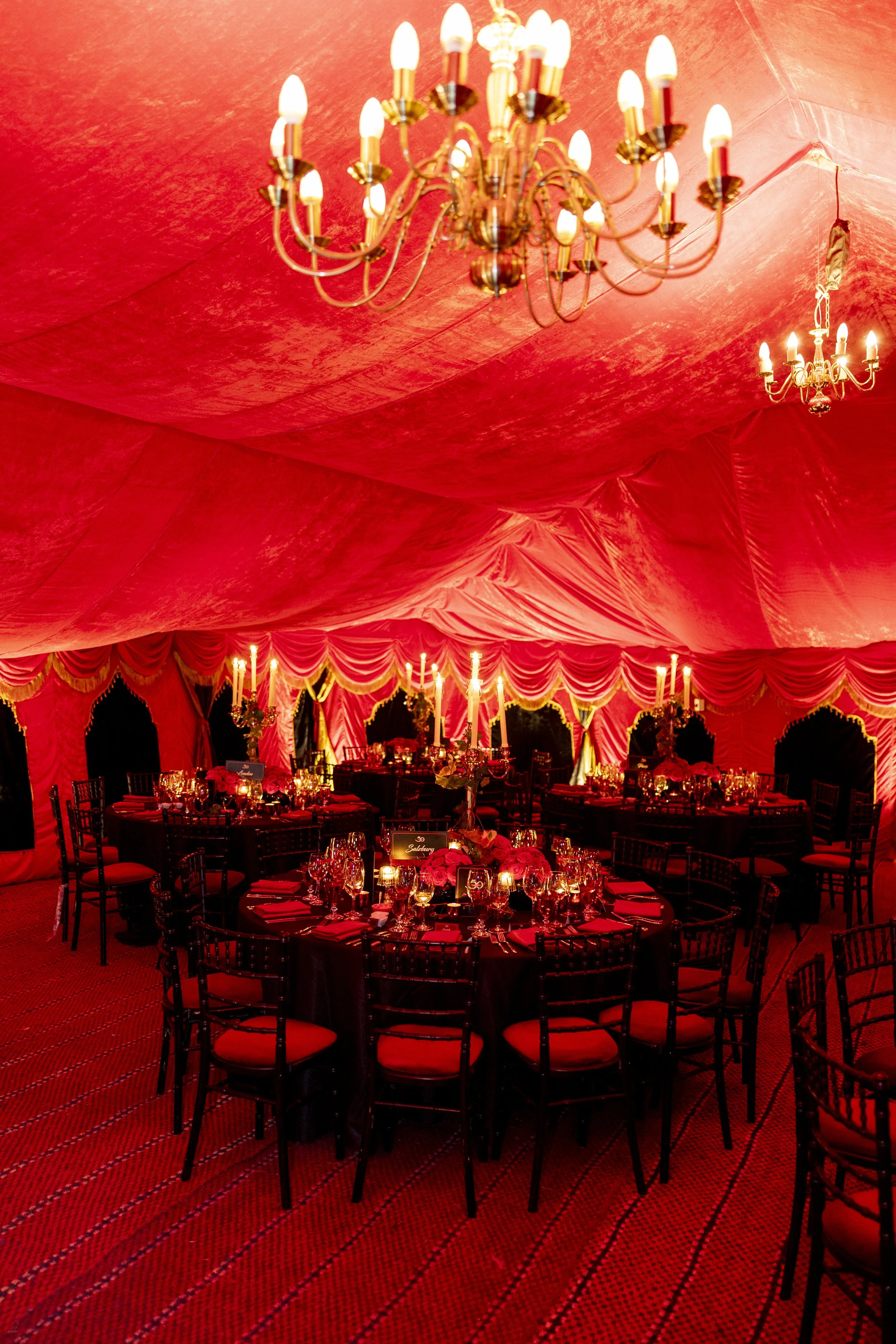 Inside a luxury red event tent with chandeliers and round tables decorated with candles, flowers, and place settings.