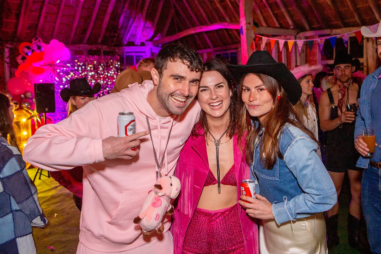 Group of friends at a party wearing western and cowboy themed outfits, smiling and holding drinks, with colorful decorations and balloons in the background.