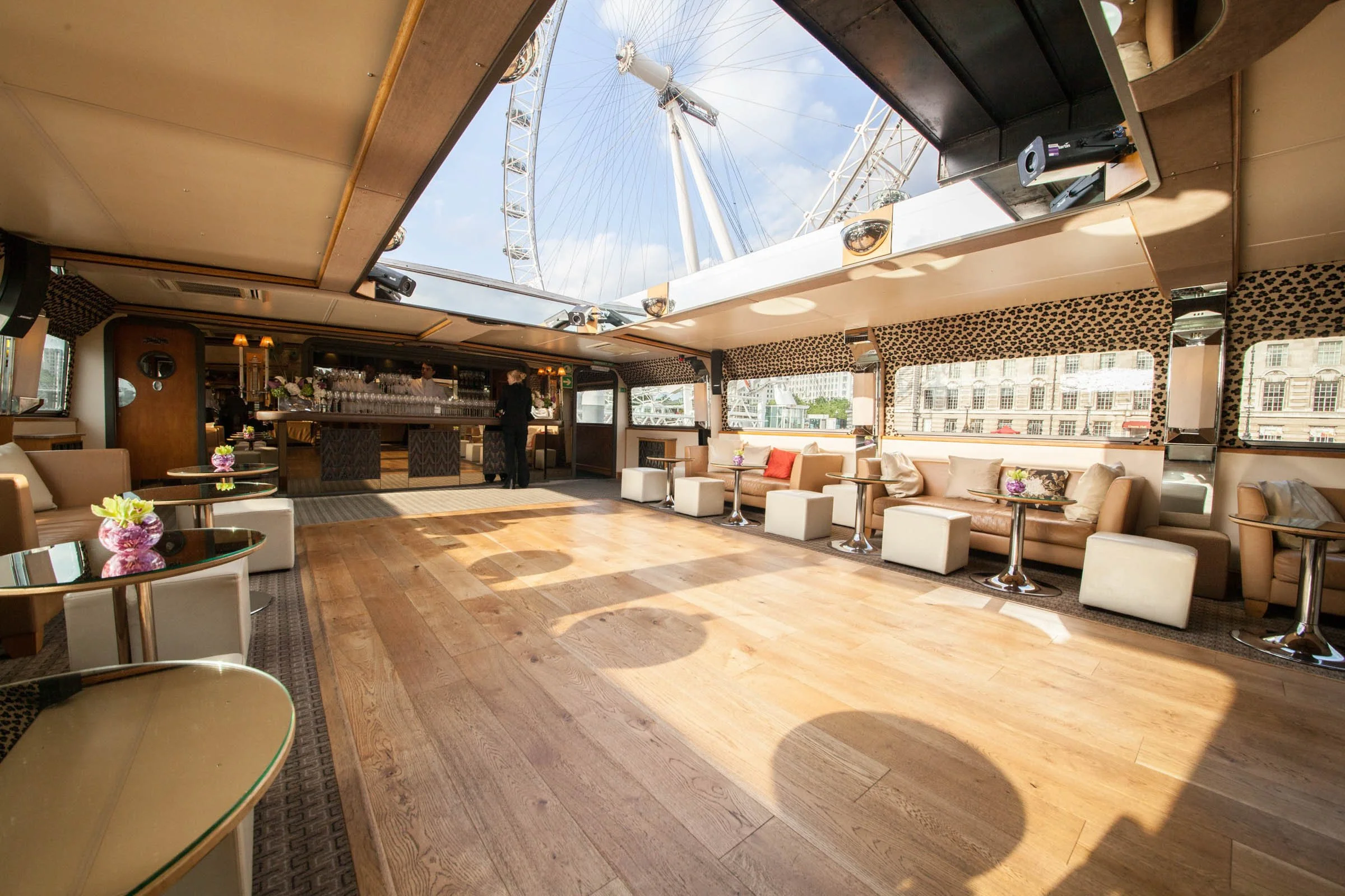Interior of a cruise ship lounge with large windows showing a Ferris wheel outside, beige seating, small tables with purple flowers, and a bar area in the background.
