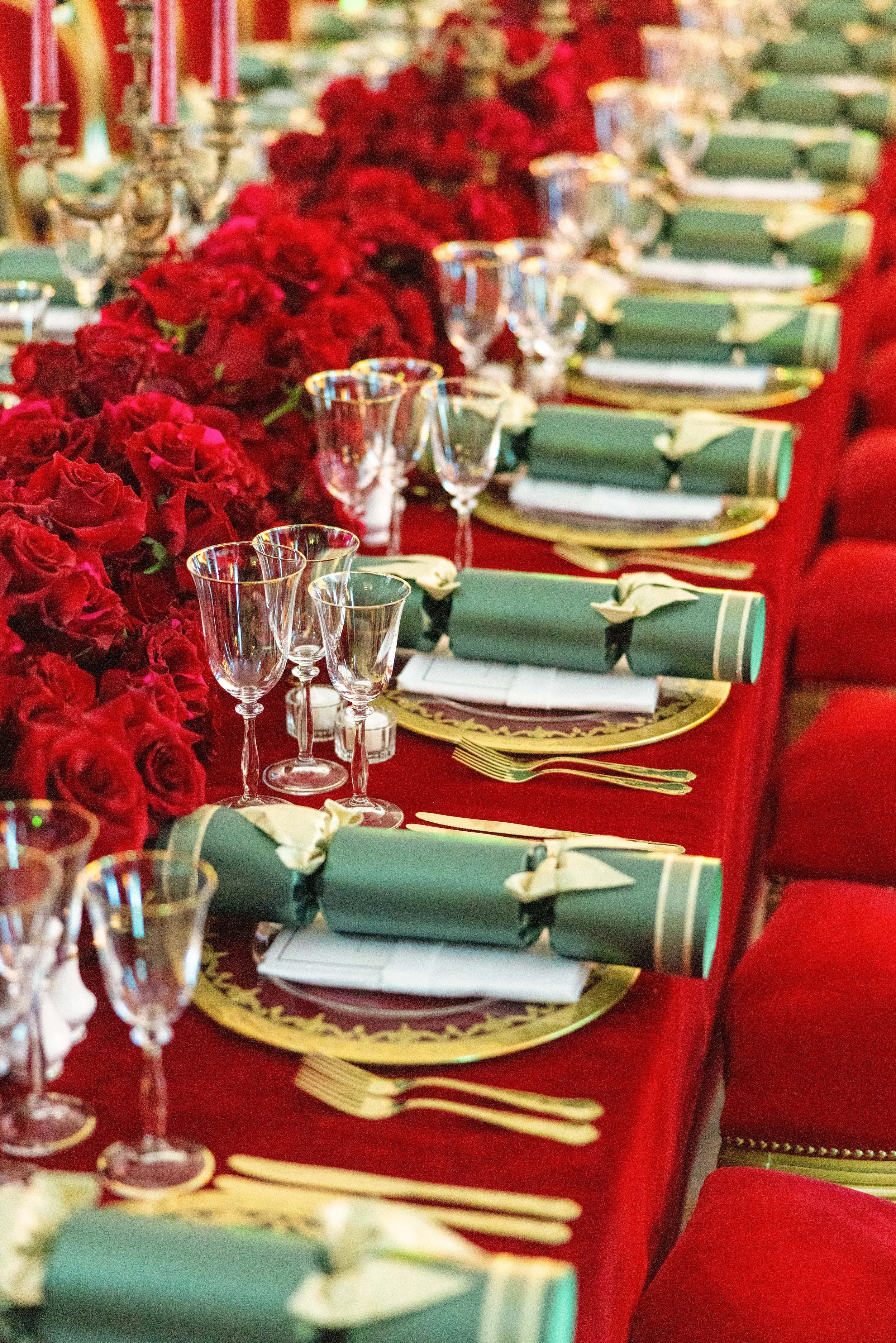 A long banquet table decorated with red roses, gold-rimmed plates, green napkins rolled with napkin rings, crystal wine glasses, and gold utensils, set for a festive event.