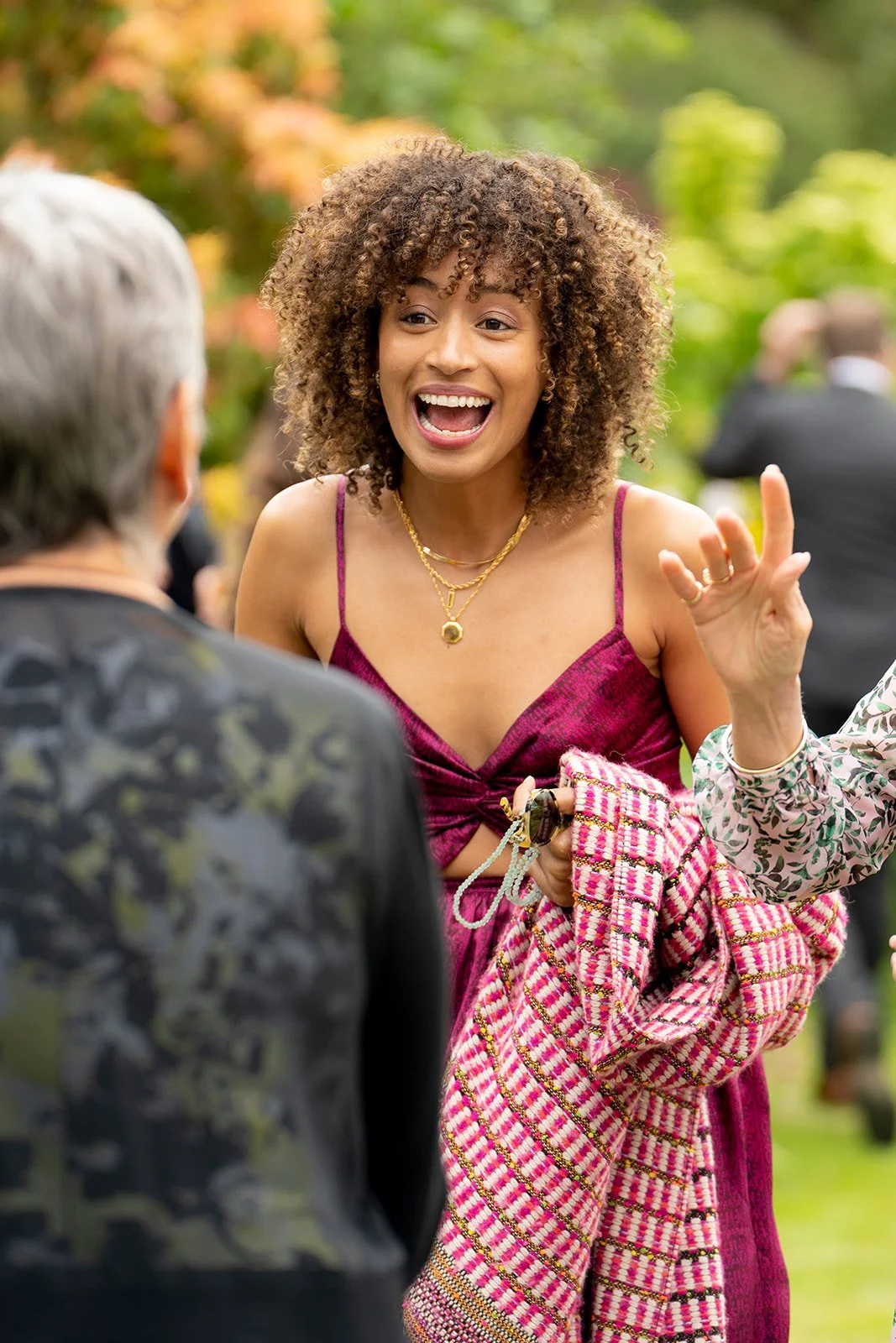 A woman with curly hair wearing a magenta dress, engaged in an animated conversation outdoors, holding a patterned scarf and smiling.