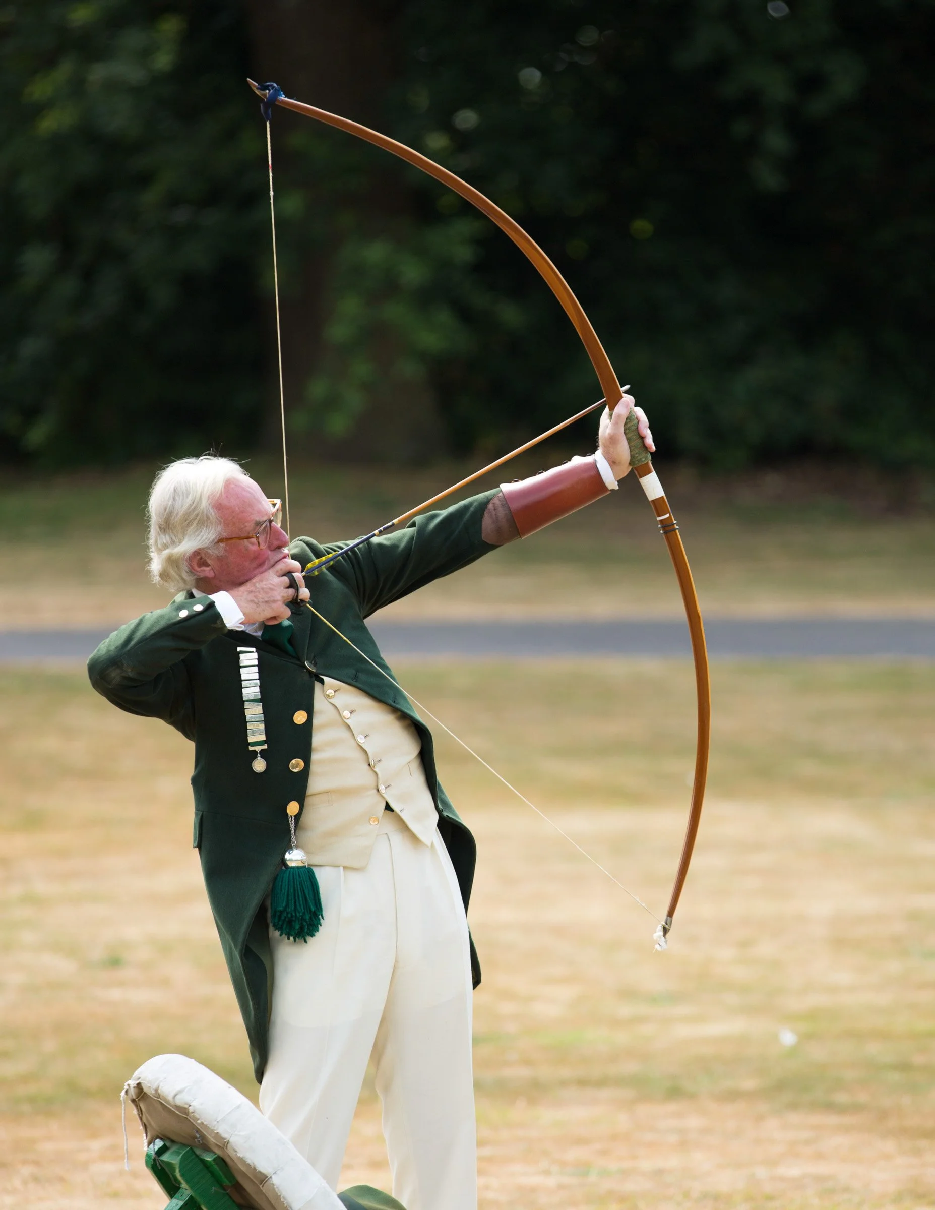 An elderly man dressed in historical clothing, aiming a bow and arrow outdoors on a grassy field with trees in the background.