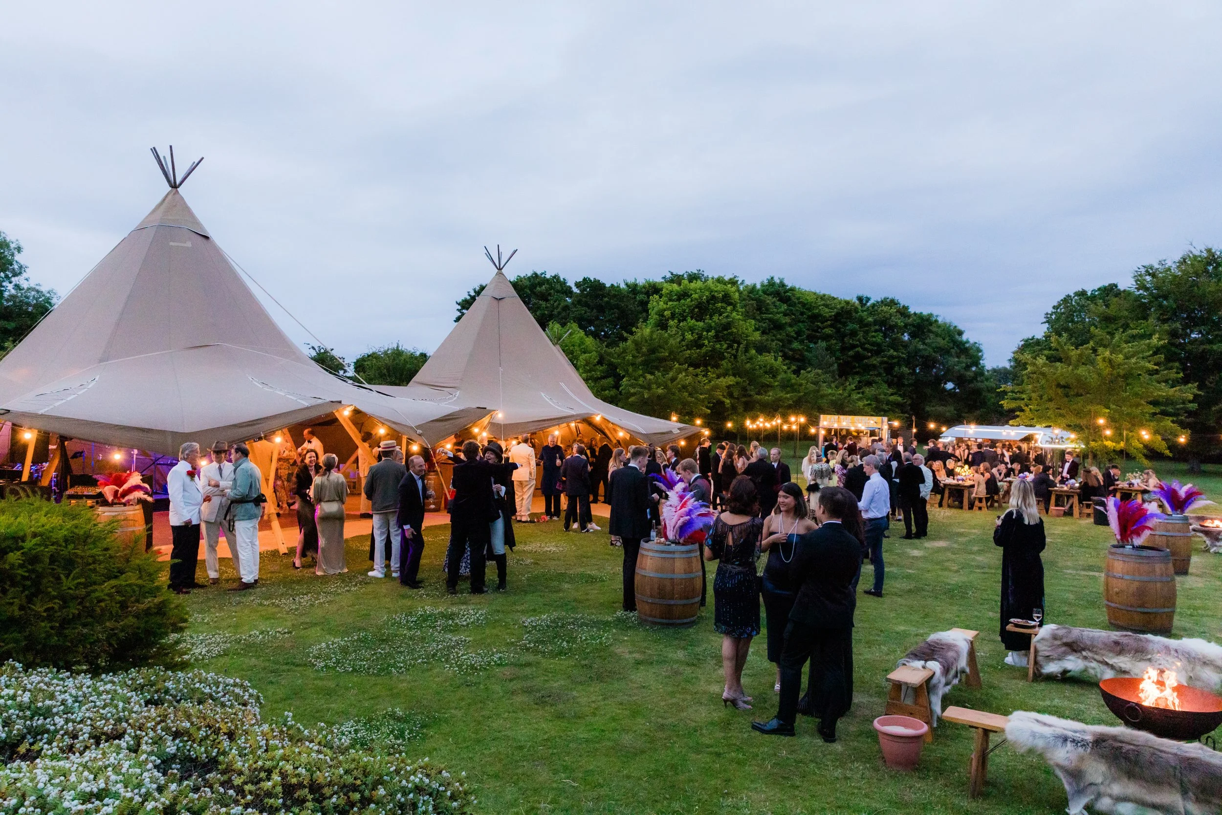 An outdoor evening event with people gathered around two large tipi tents decorated with string lights, surrounded by trees and greenery.