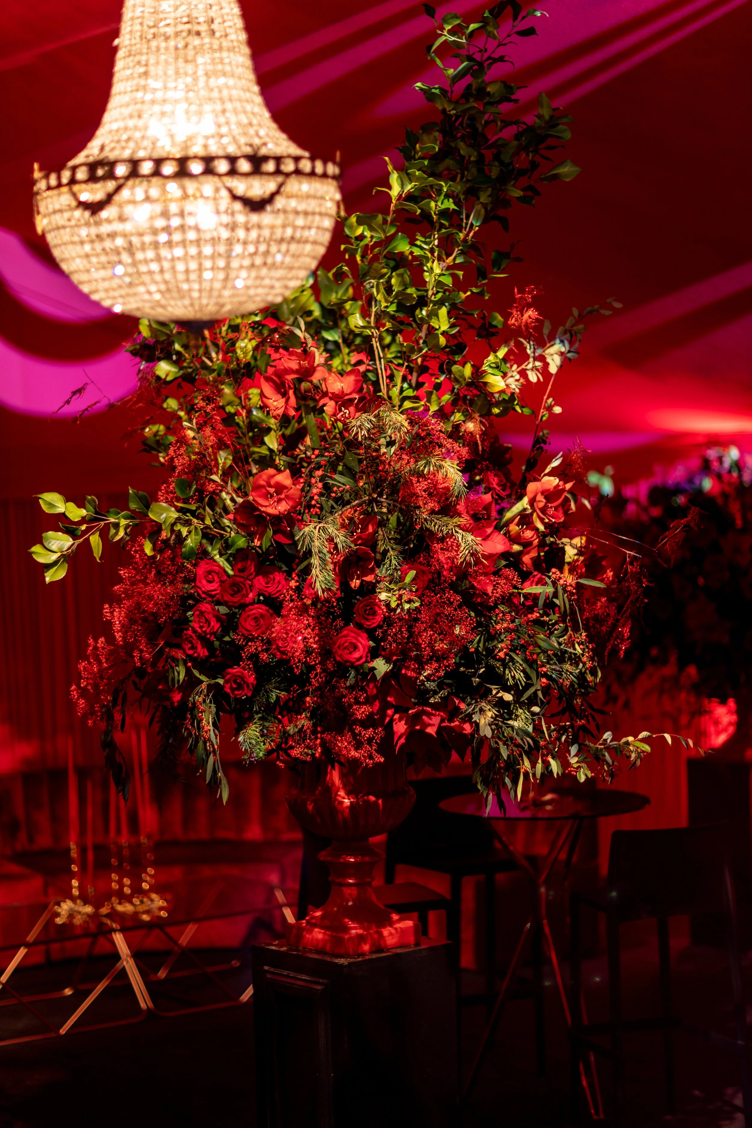 A large floral arrangement of red roses, red flowers, and greenery in a decorative vase, placed on a black pedestal, with a chandelier hanging above, set in a dimly lit room with red lighting.