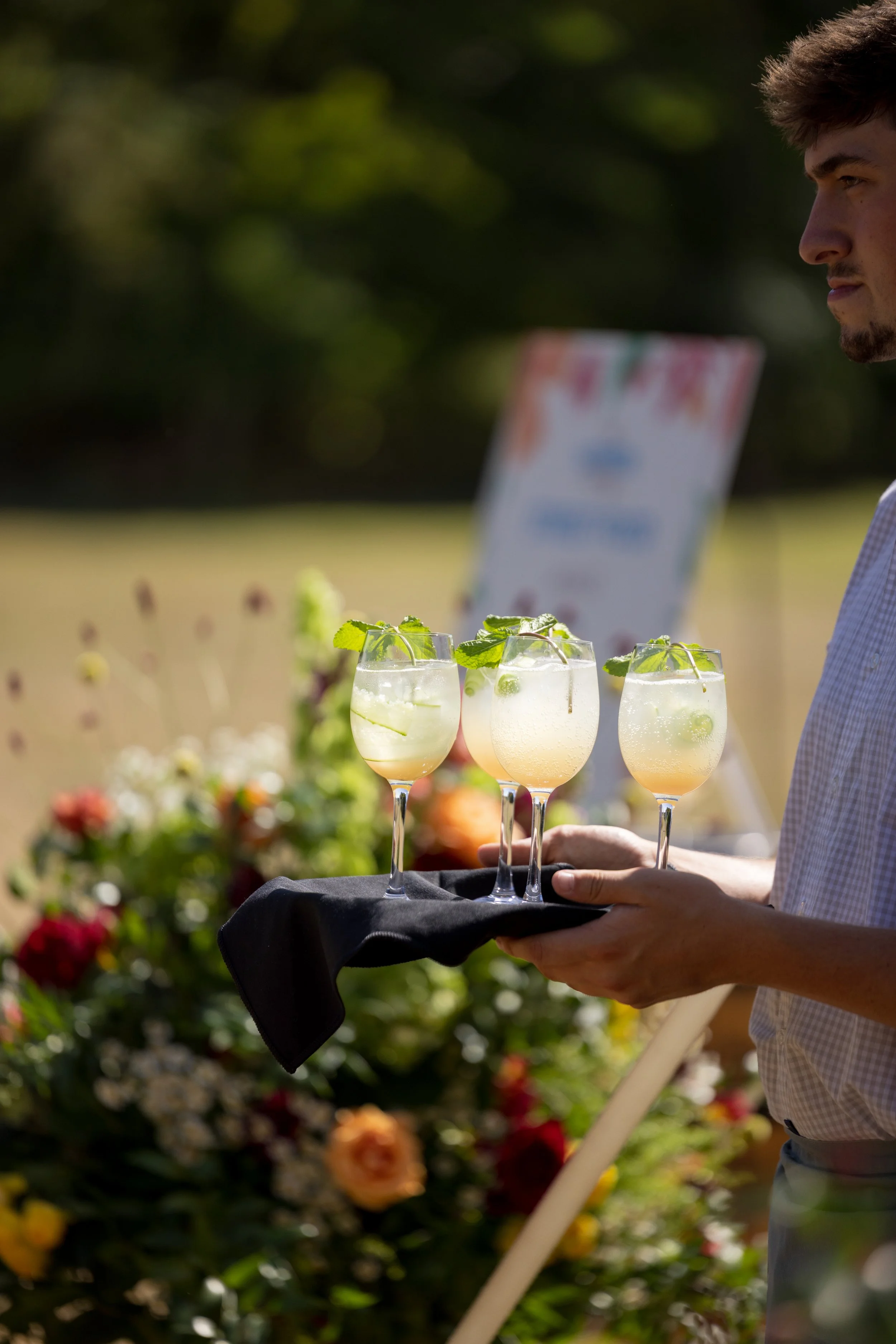 A person holding a tray with three glasses of a lemon-lime cocktail garnished with mint leaves at an outdoor event with a flower display in the background.