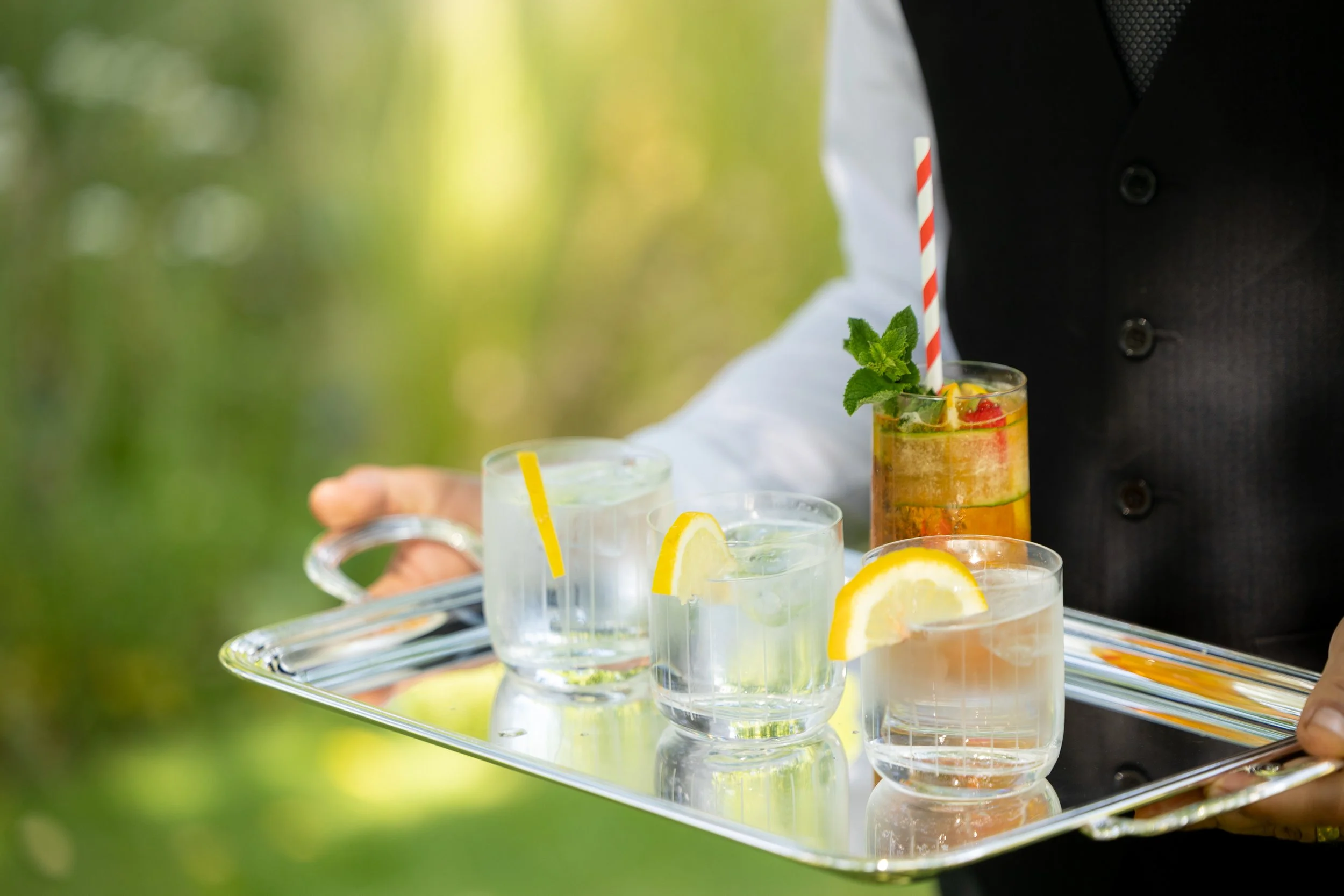 A person in formal attire holds a silver tray with four drinks, three of which are lemonade with lemon wedges, and one is a colorful cocktail garnished with mint, straw, and fruit, outdoors with a blurred green background.