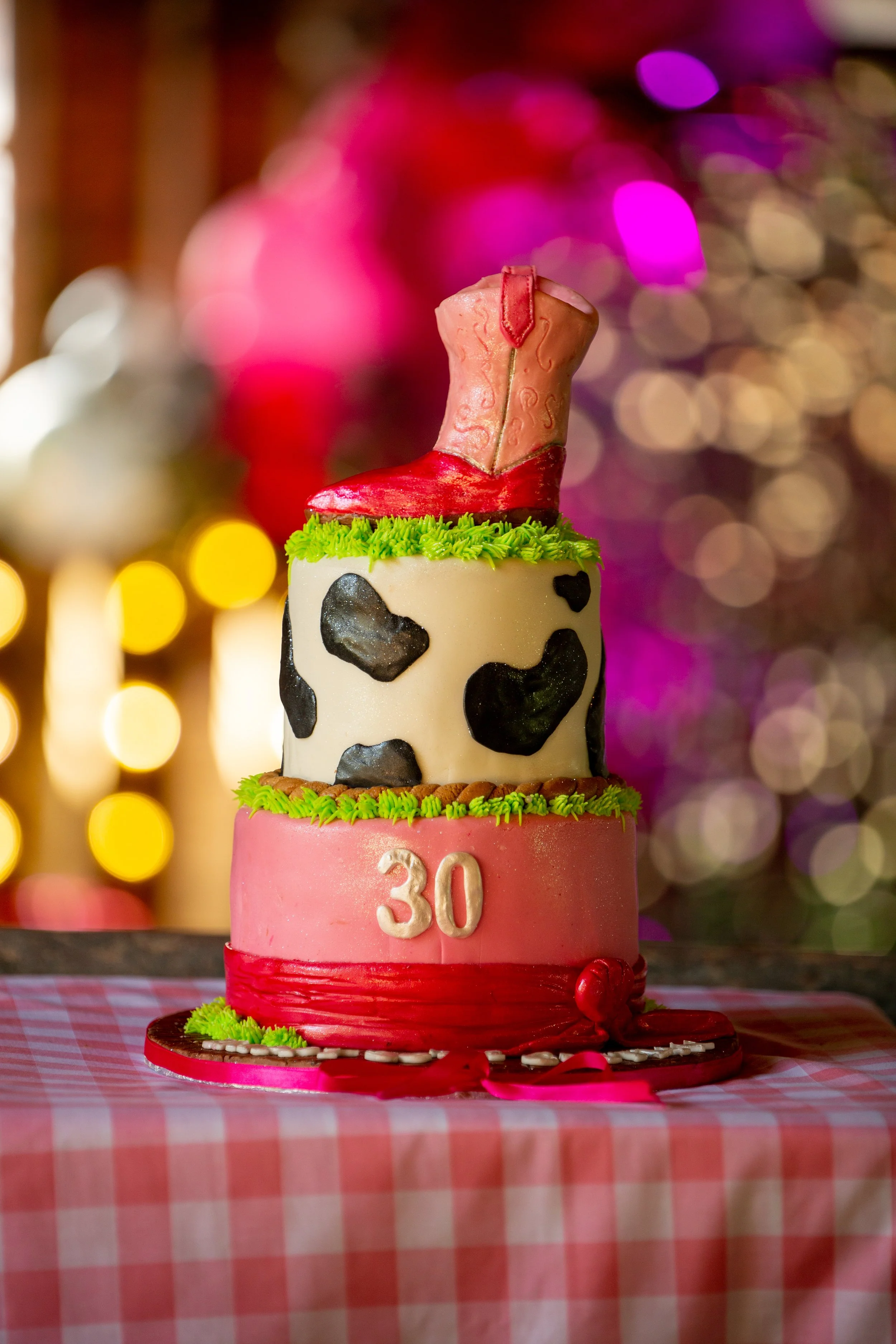 A three-tier birthday cake with cow print, pink, and a cowboy boot topper on a checkered tablecloth with festive, colorful background lights.