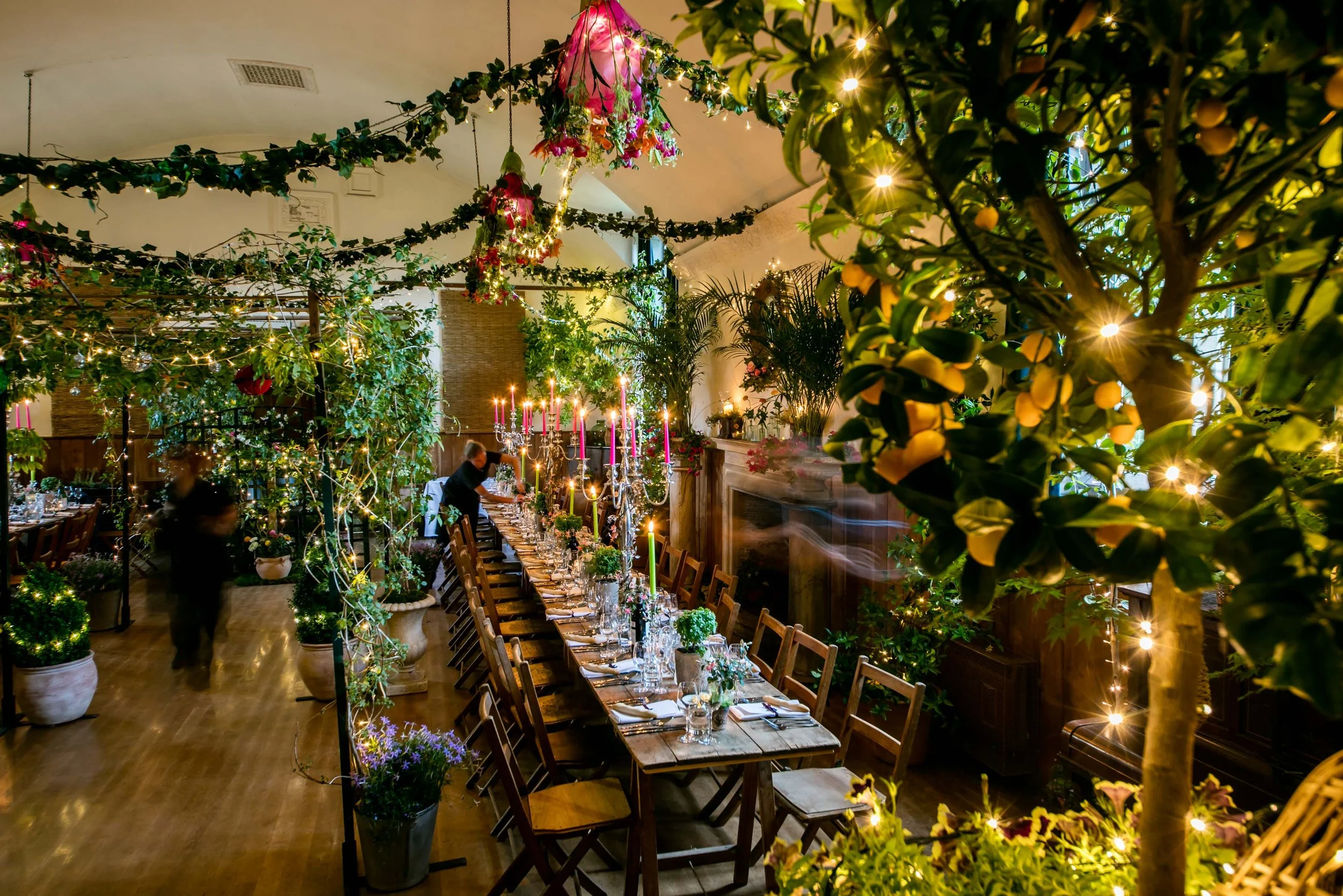 A long dining table set with glassware, candles, and floral centerpieces in a decorated indoor space with string lights, greenery, and hanging flowers.
