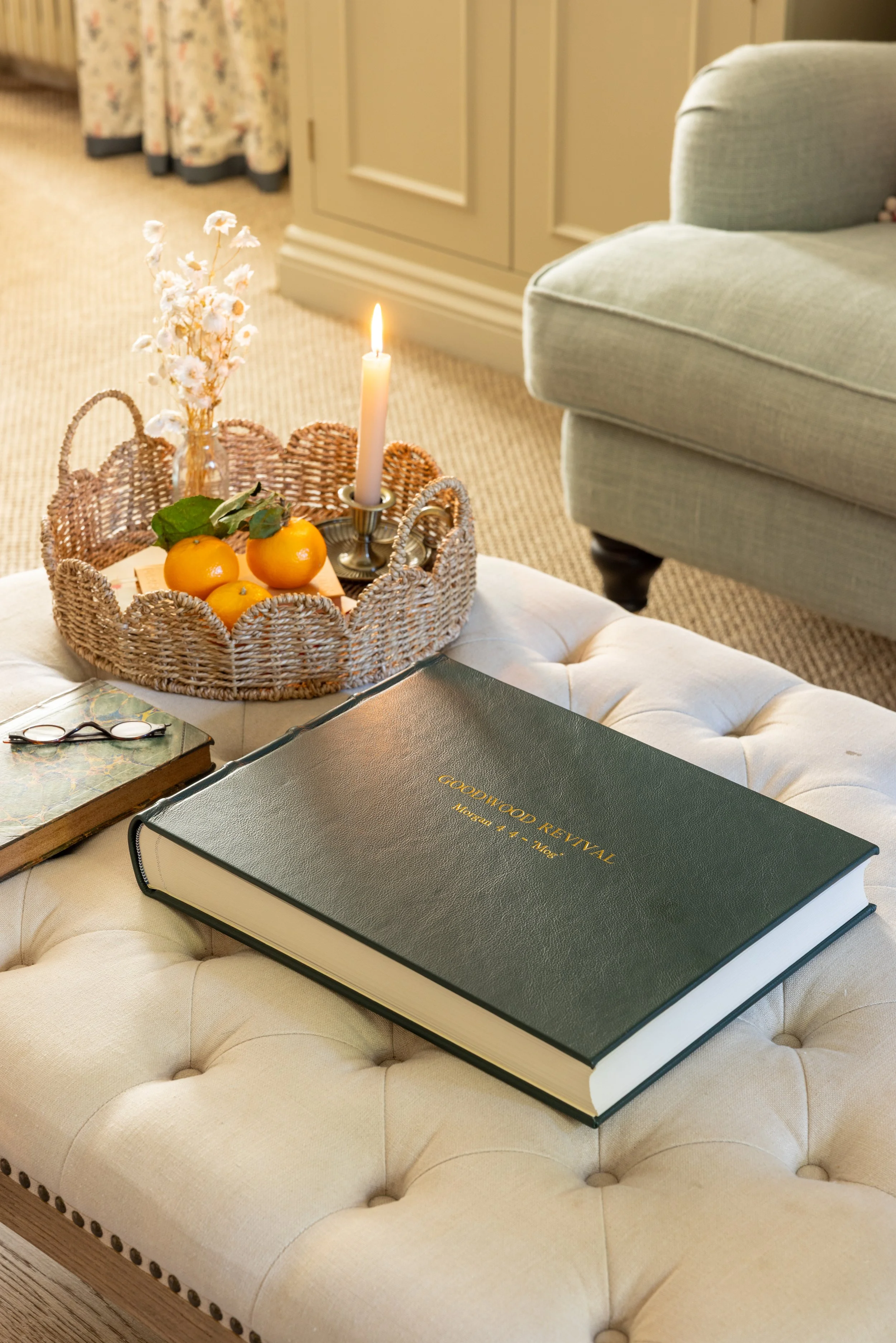 A beige upholstered tufted ottoman with a black hardcover book titled 'Groomed Revival' placed on it. On the book are eyeglasses. Next to the book, there is a round wicker tray with three oranges, a small plant, a white candle, and a glass vase with 