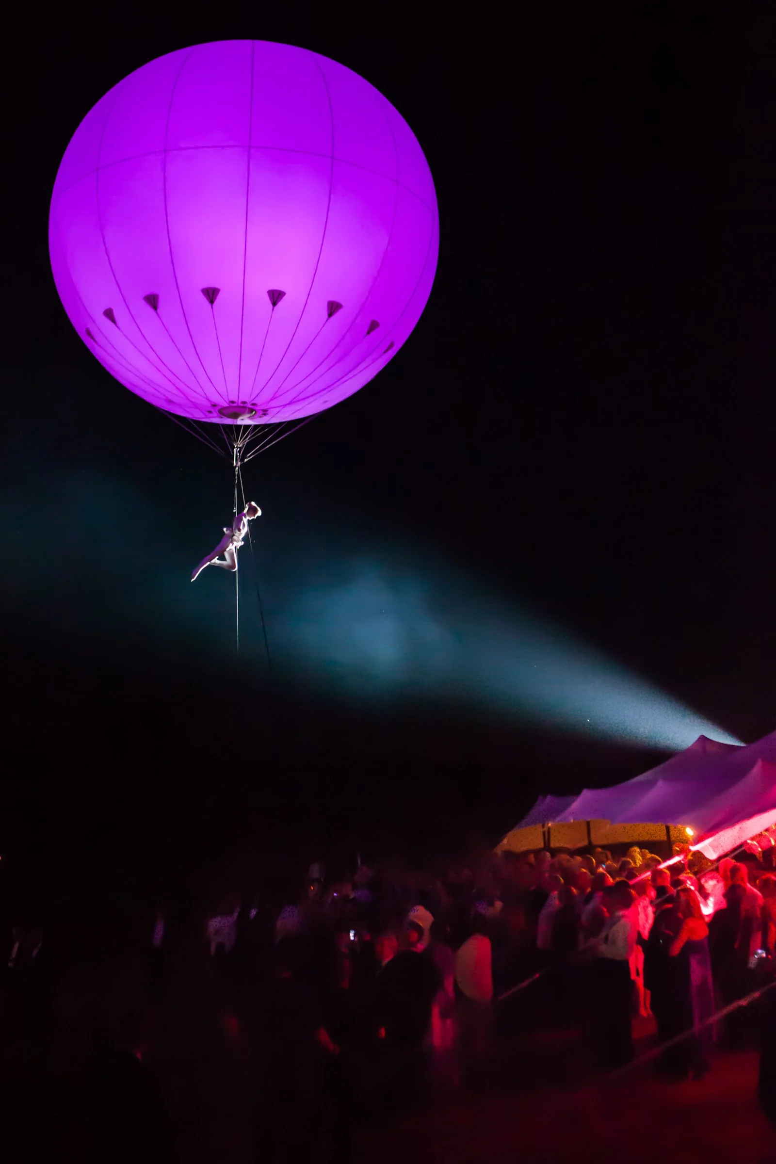 A performer hanging from a large purple illuminated balloon at night during a public event, with a crowd of people and tents below.