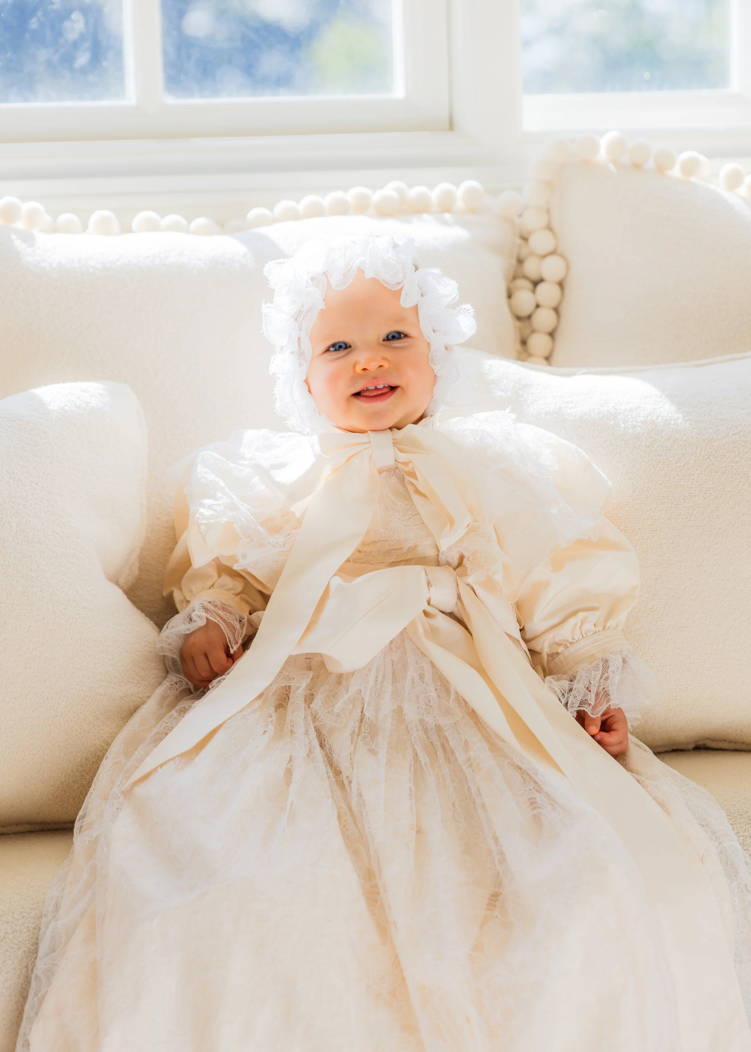 A smiling baby girl sitting on a cream-colored couch in a lace and satin dress, with a matching bonnet, in front of a window.