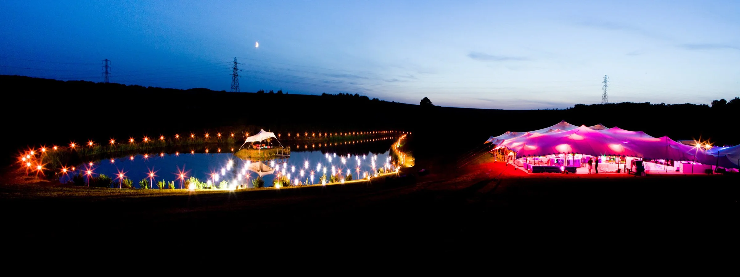 Night scene with illuminated outdoor event, featuring a large pink and purple tent and a small illuminated pond with surrounding lights and a tent with a white canopy.