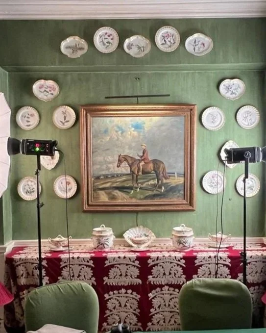 A room with green walls decorated with decorative plates arranged around a framed painting of a person riding a horse, with two studio lights on stands, and a table draped with a red tablecloth with white patterns and two decorative jars.