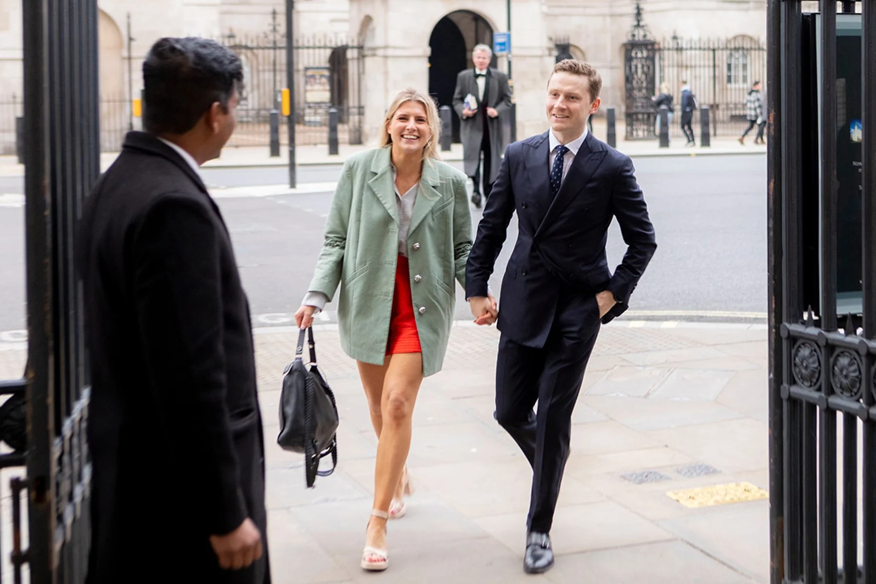A woman in a red skirt and green coat, and a man in a dark suit, are walking out of a building, smiling and holding hands. A person in a suit is standing nearby.
