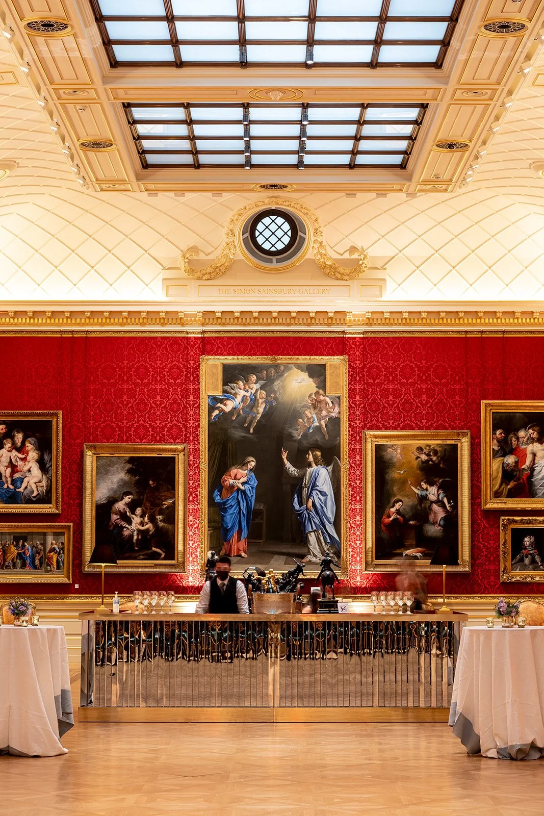 Interior of an art gallery with ornate decoration, red damask wallpaper, classical paintings, and a mirrored reception desk in front of wall-mounted artworks.