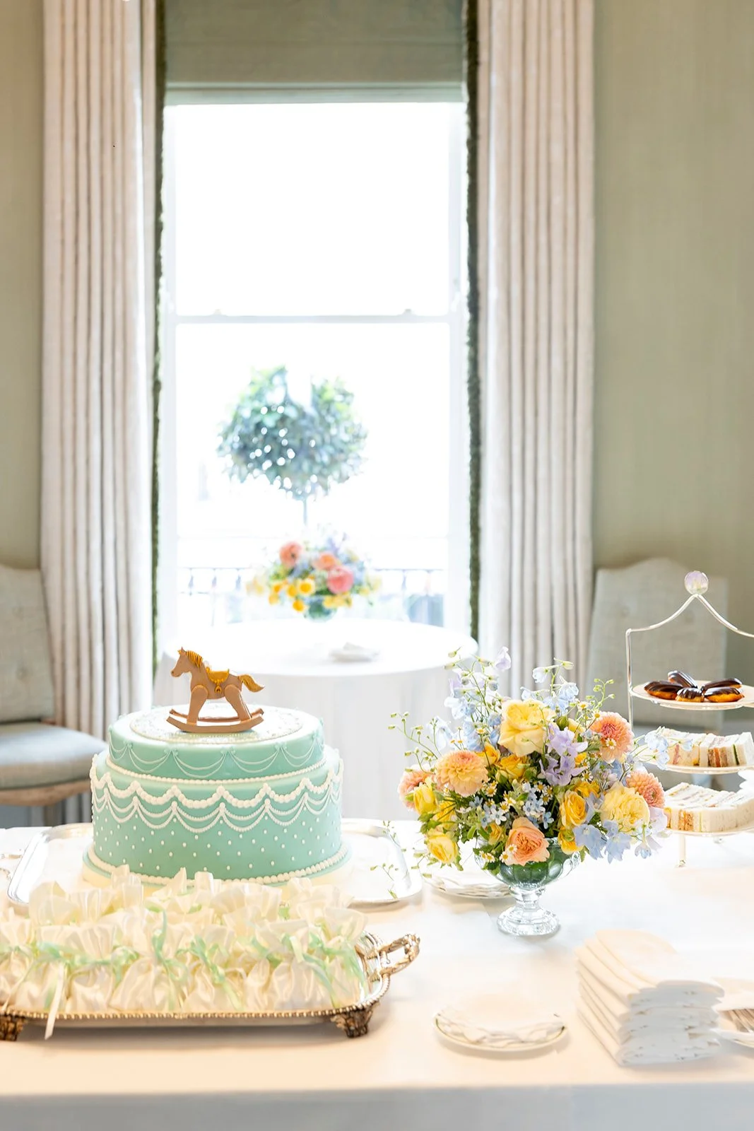 Cake with a rocking horse topper, floral arrangement, and sweets on a table set for a celebration in a bright room.