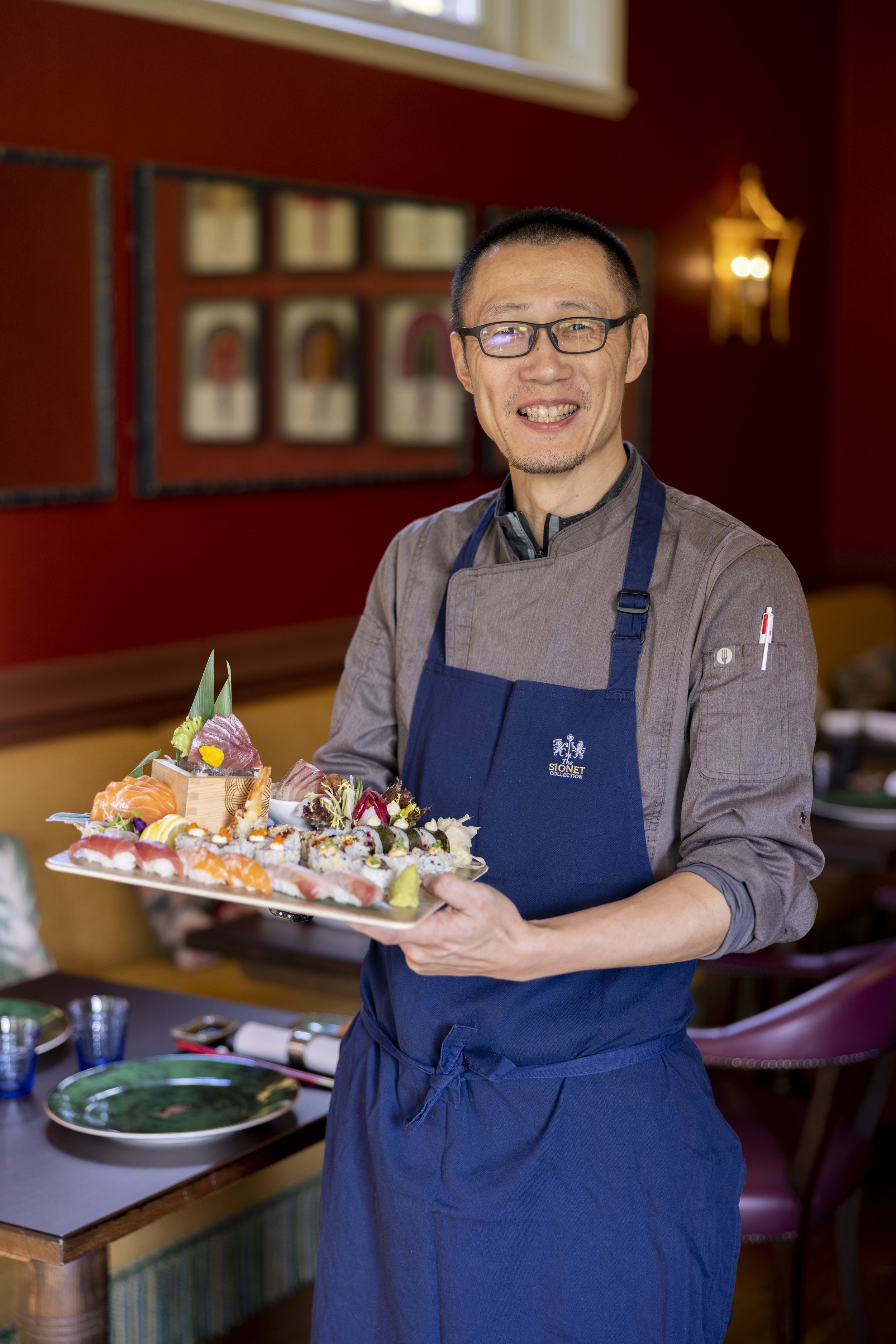 A chef holding a tray of assorted sushi in a restaurant with red walls and framed pictures in the background.