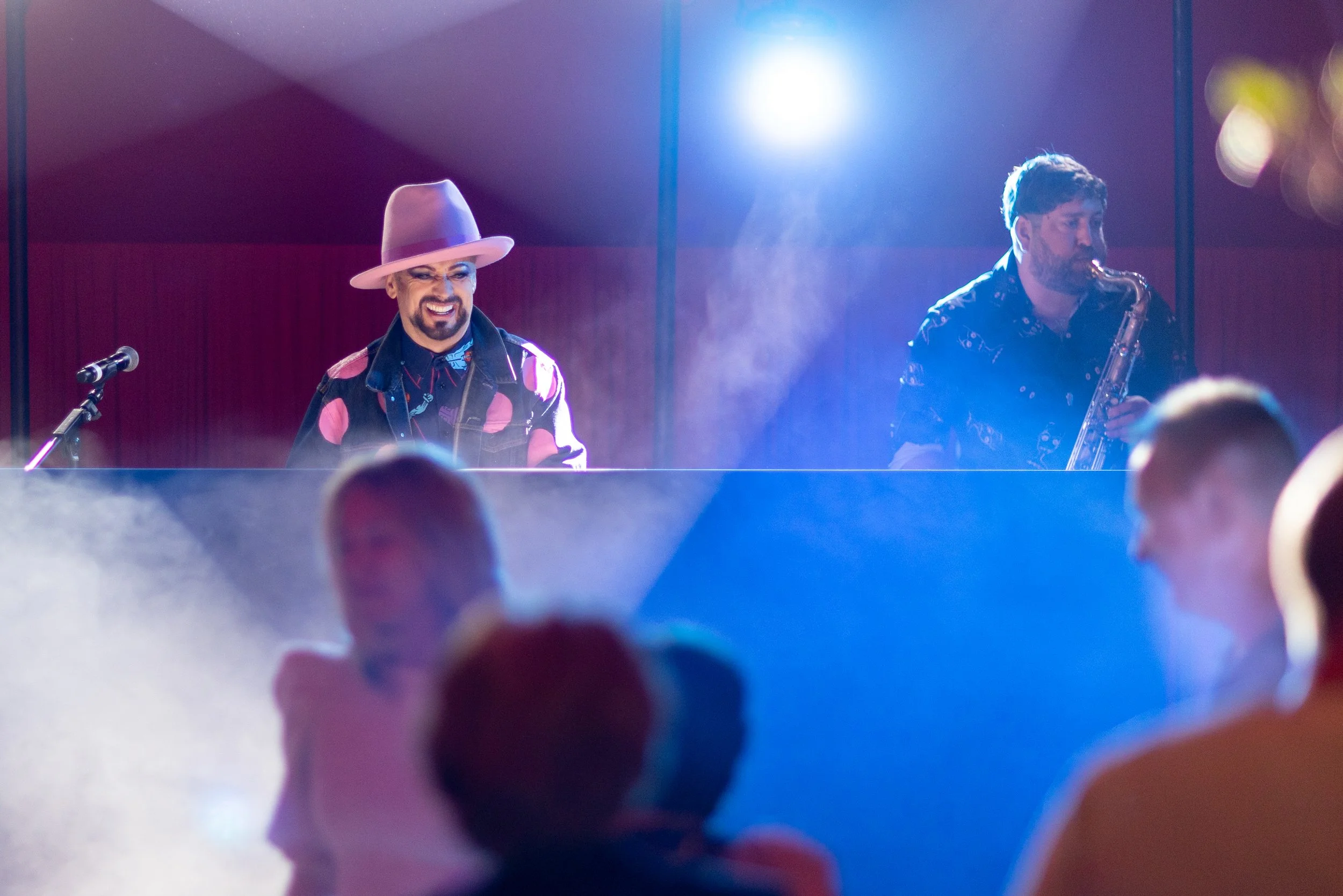 A musical performance featuring a smiling man wearing a pink wide-brimmed hat and colorful shirt playing the piano, alongside a man playing a saxophone on stage, with audience members watching.