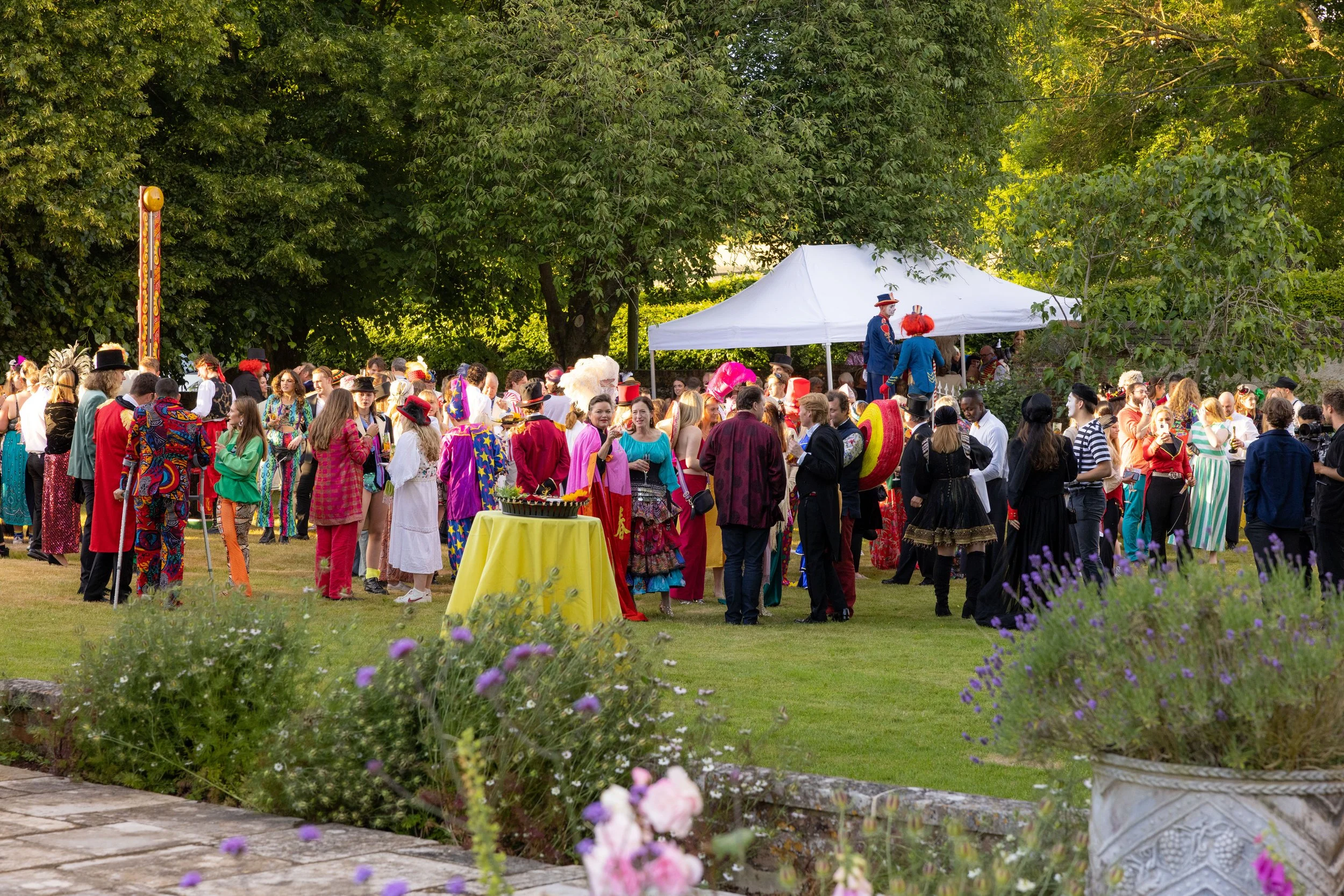 People dressed in colorful and vintage costumes gathered outdoors for a celebration or festival under a large tree with a white canopy in the background.