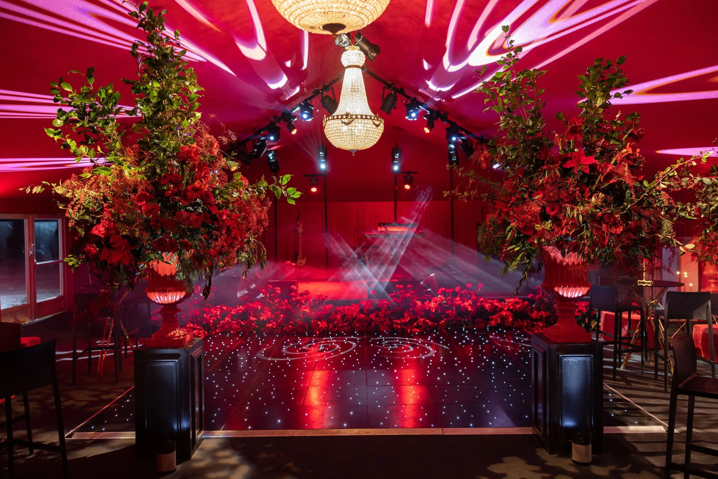 A decorated event space with a red theme, featuring large floral arrangements, a chandelier, dance floor with twinkling lights, stage with lighting and a grand piano, and tables with chairs for a celebration or party.