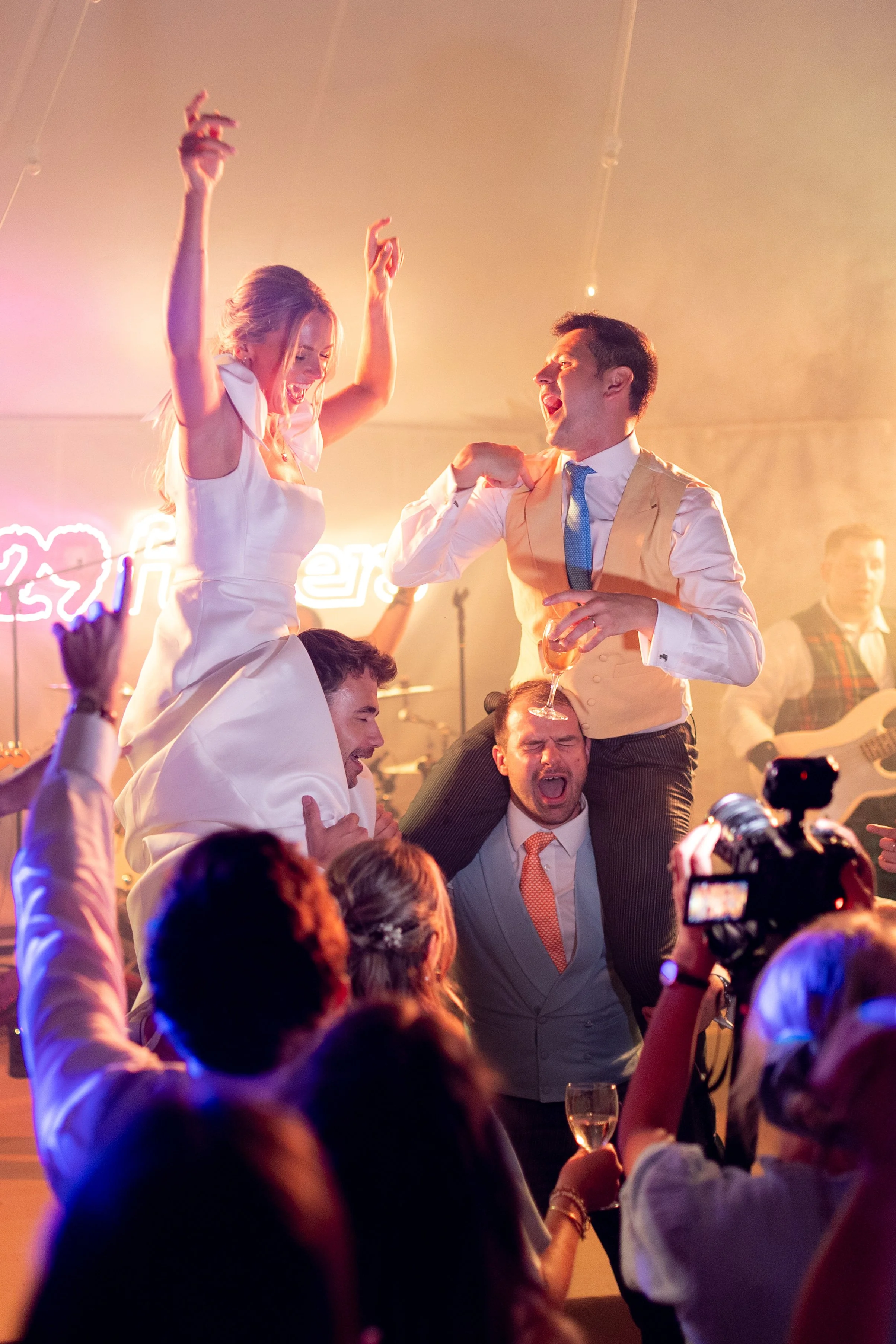 People celebrating at a lively party or event, with some individuals dancing and others holding drinks, in a warmly lit room.
