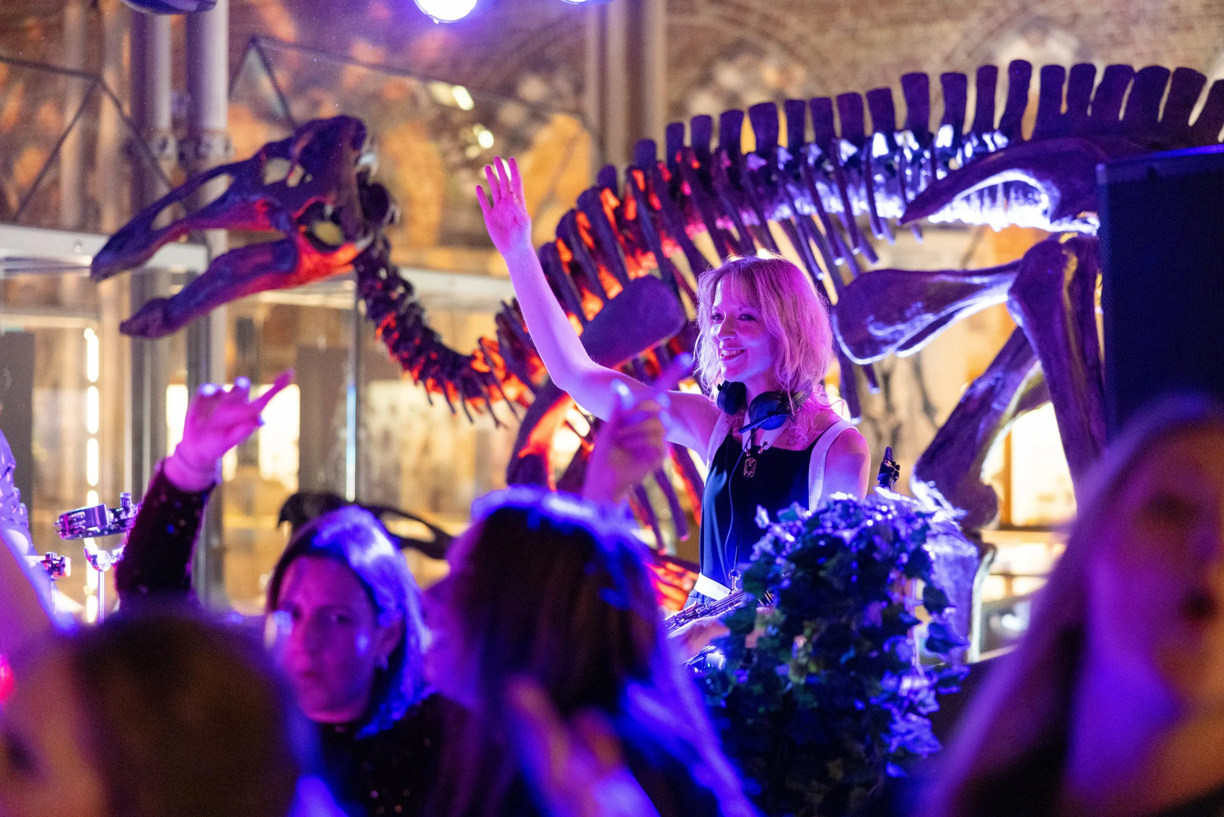 A woman with blonde hair and headphones DJing at a lively event, surrounded by people, with a large dinosaur skeleton illuminated in the background.