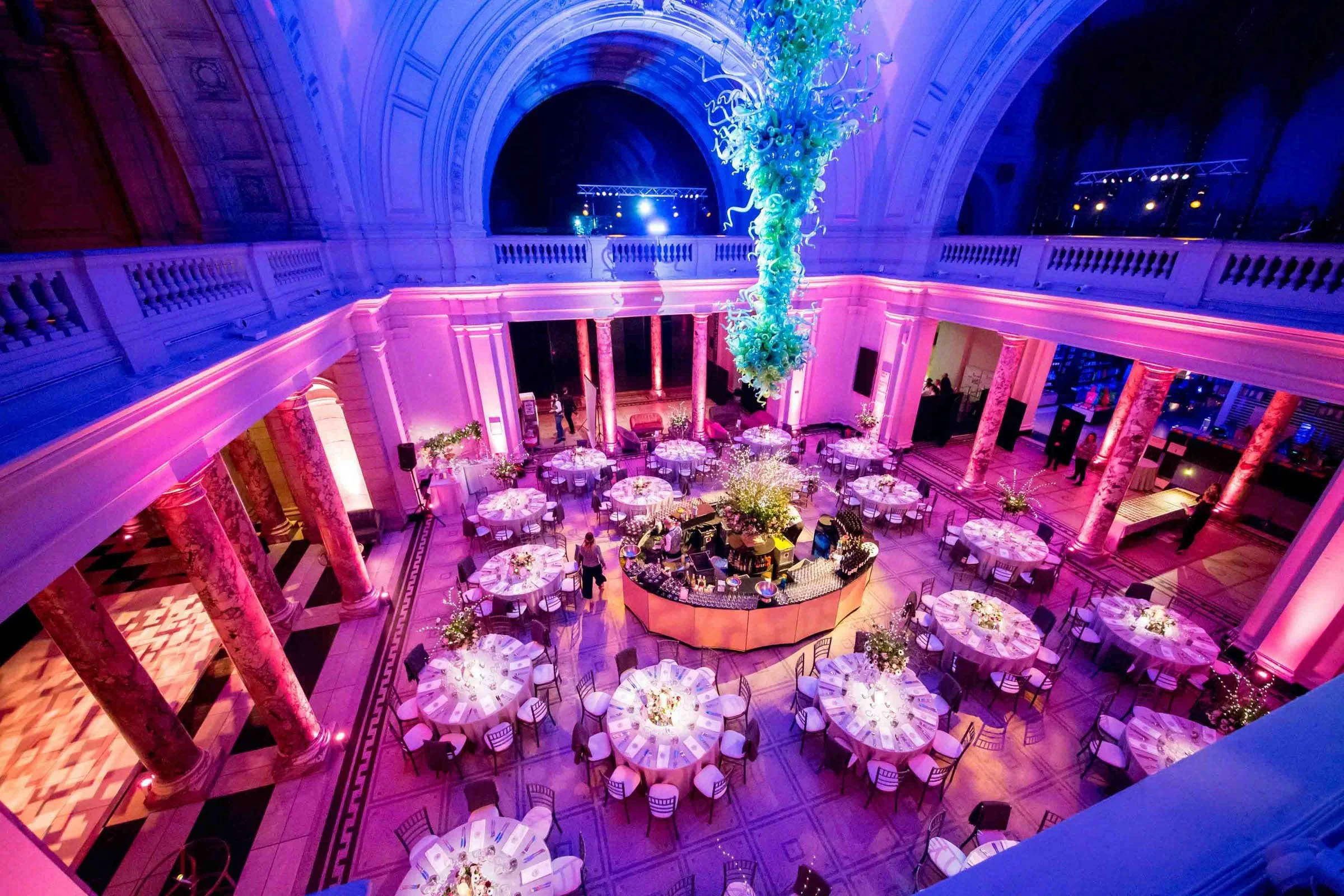 A grand ballroom decorated for a formal event with round tables, floral centerpieces, pink and purple lighting, and a large hanging glass sculpture in the center.