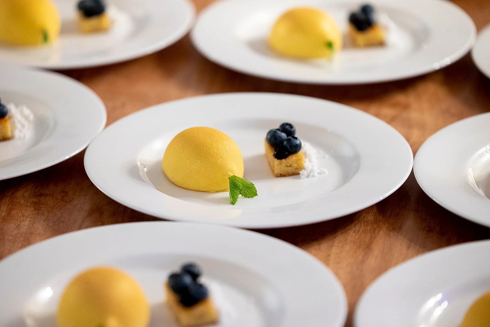 Multiple white plates with yellow dome-shaped desserts, small square pastries topped with blueberries, and a green mint leaf on each plate, arranged on a wooden surface.