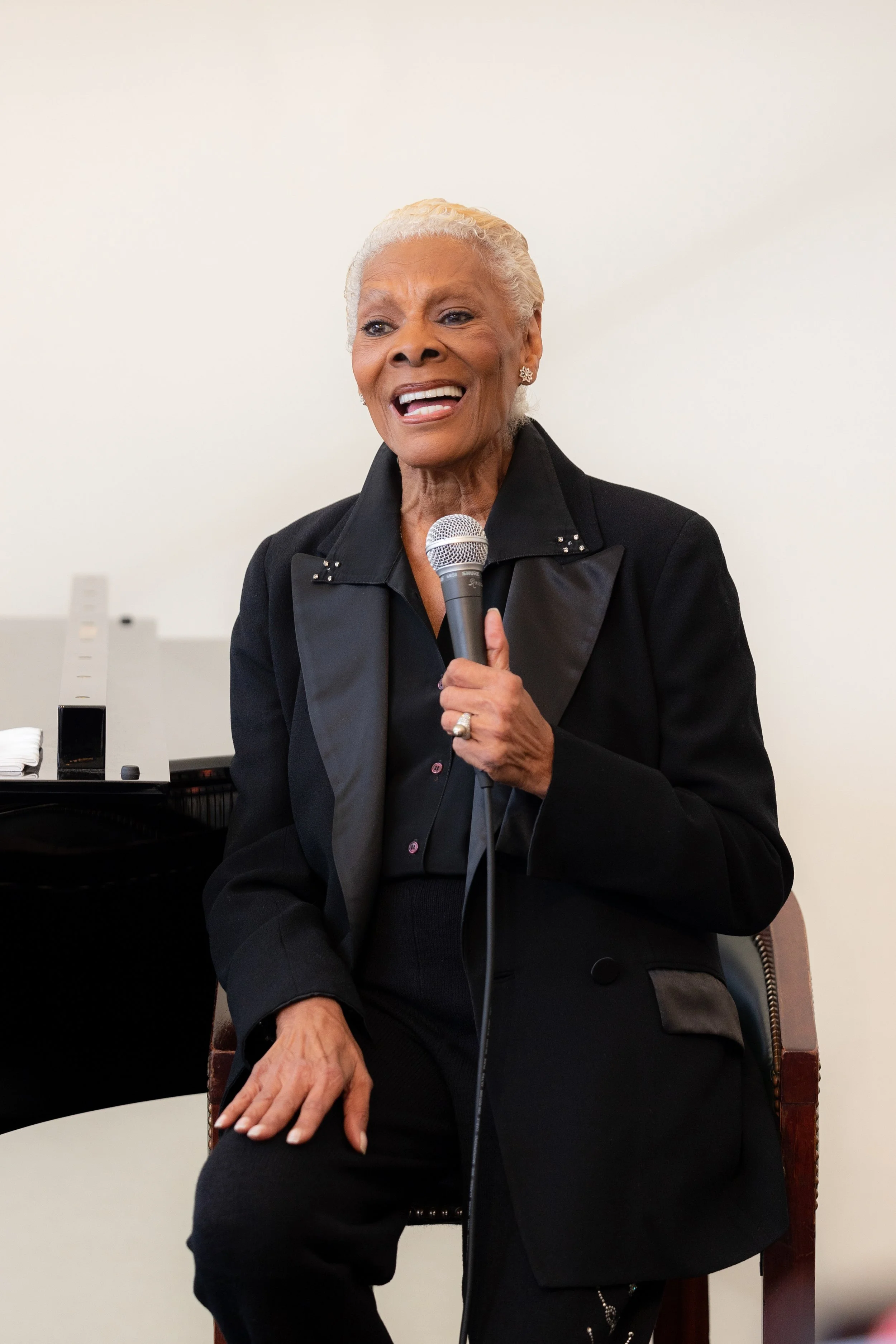 An elderly woman with white hair is speaking into a microphone while sitting on a chair. She is wearing a black suit with a satin lapel and has a happy expression.