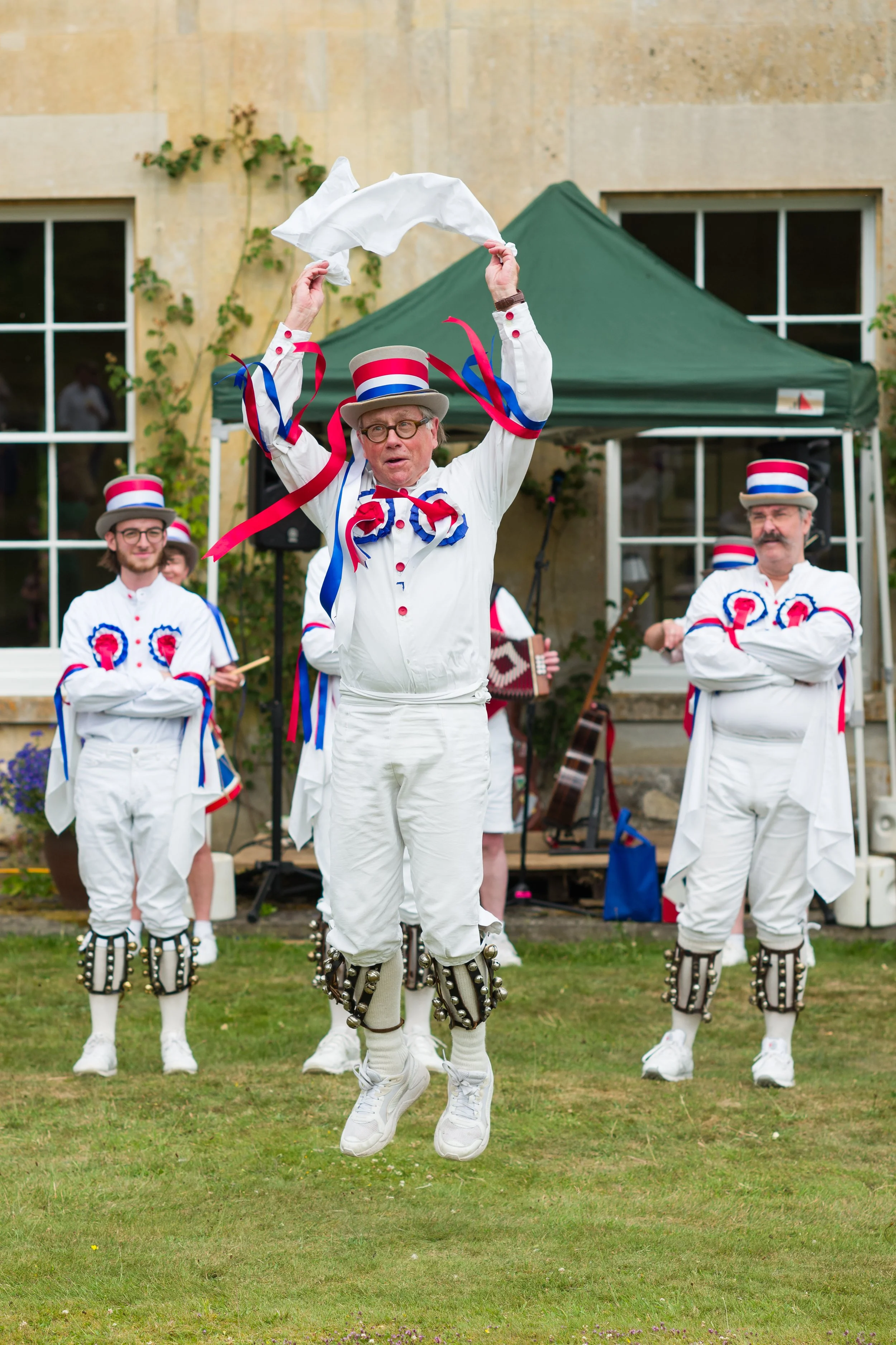 Men dressed in patriotic costumes performing a traditional dance outdoors, with a green tent and a beige building in the background.