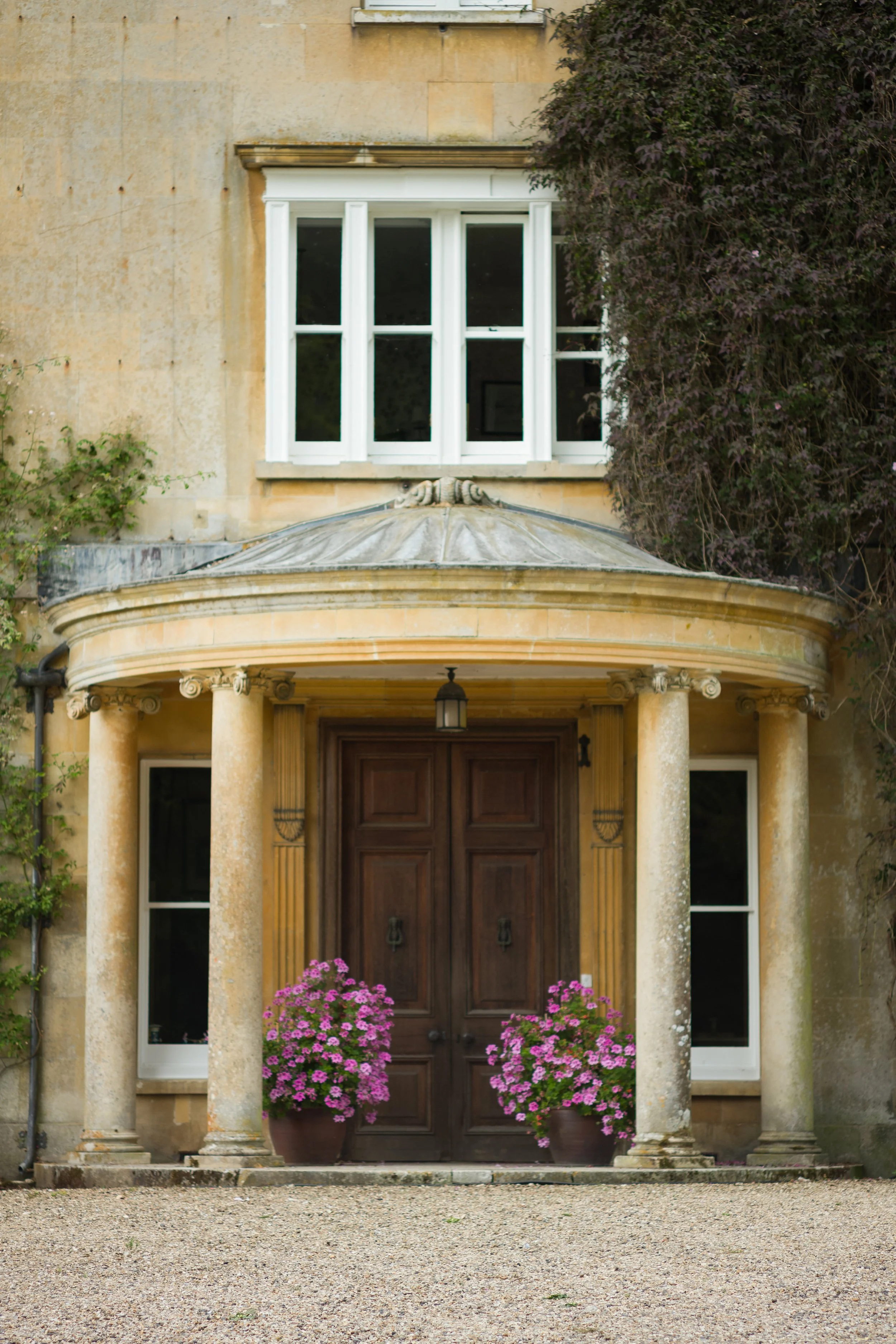 The front entrance of an elegant house with a large wooden double door, framed by two classical columns, two pink flower pots, and a portico canopy. There are windows above and beside the door.