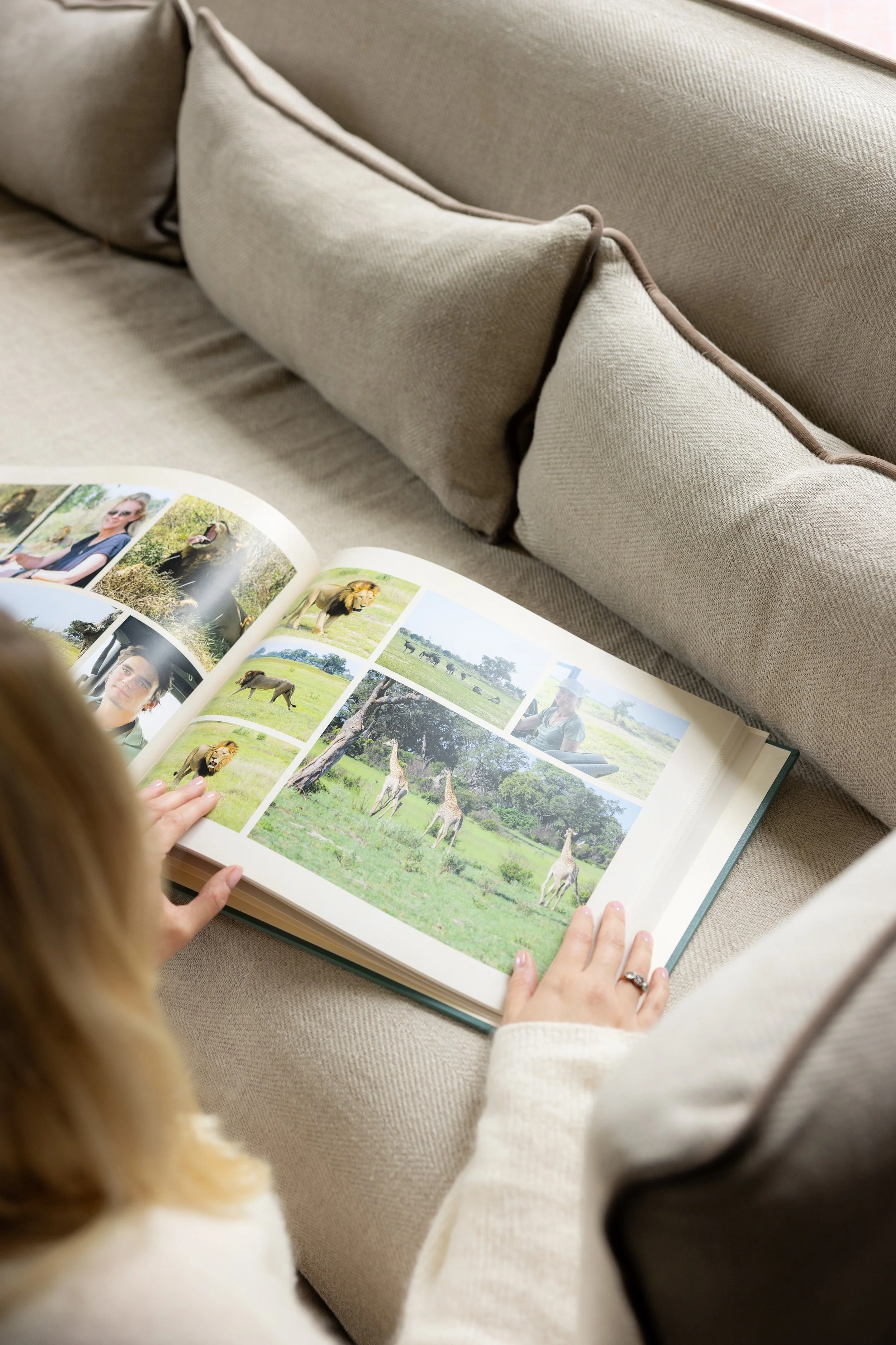 A woman sitting on a beige couch looking at a photo album with pictures of lions, giraffes, and people in a safari setting.