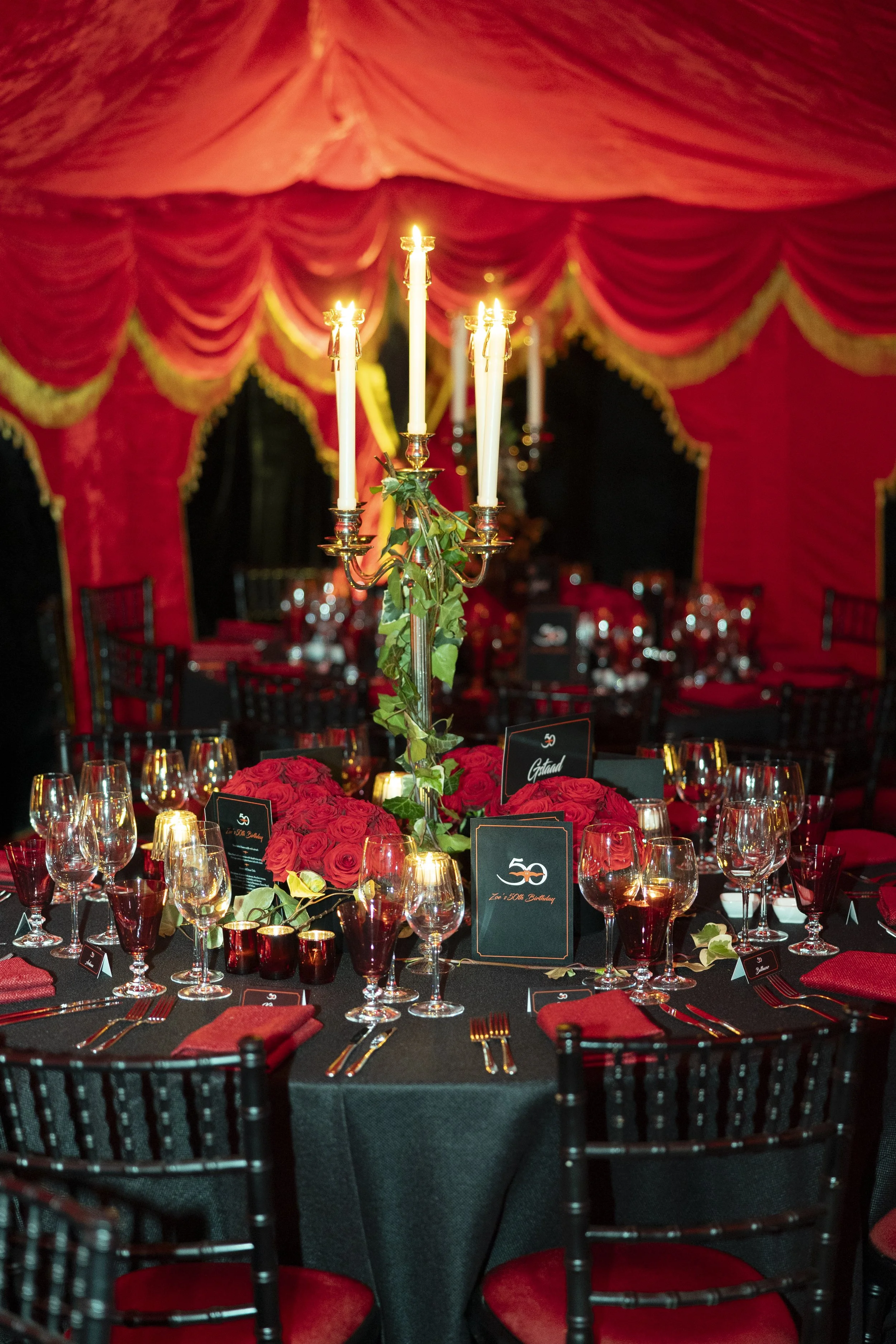 A formal banquet table decorated with red roses, candles, and elegant glassware inside a red and gold draped tent.