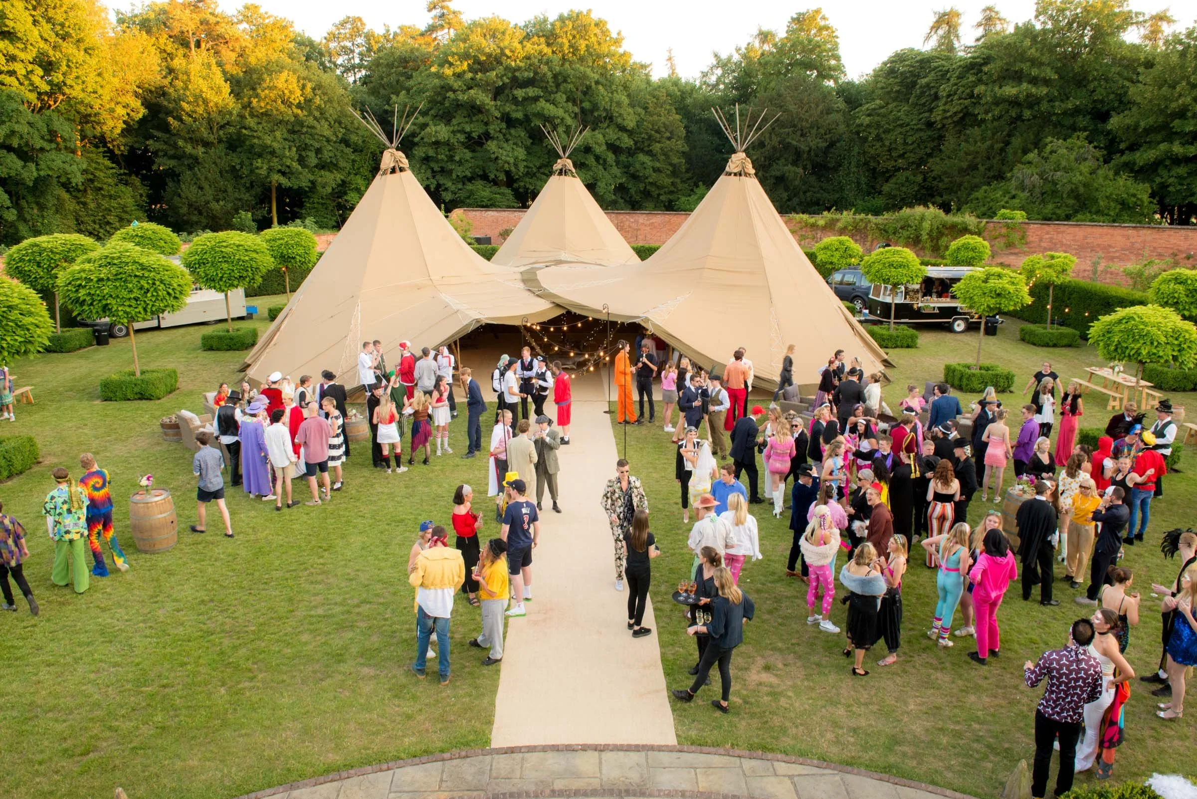 Outdoor party scene with a large beige teepee tent, decorated with string lights, surrounded by people dressed in colorful and vintage clothing. There are small round trees and a brick wall in the background, and food trucks are parked nearby.