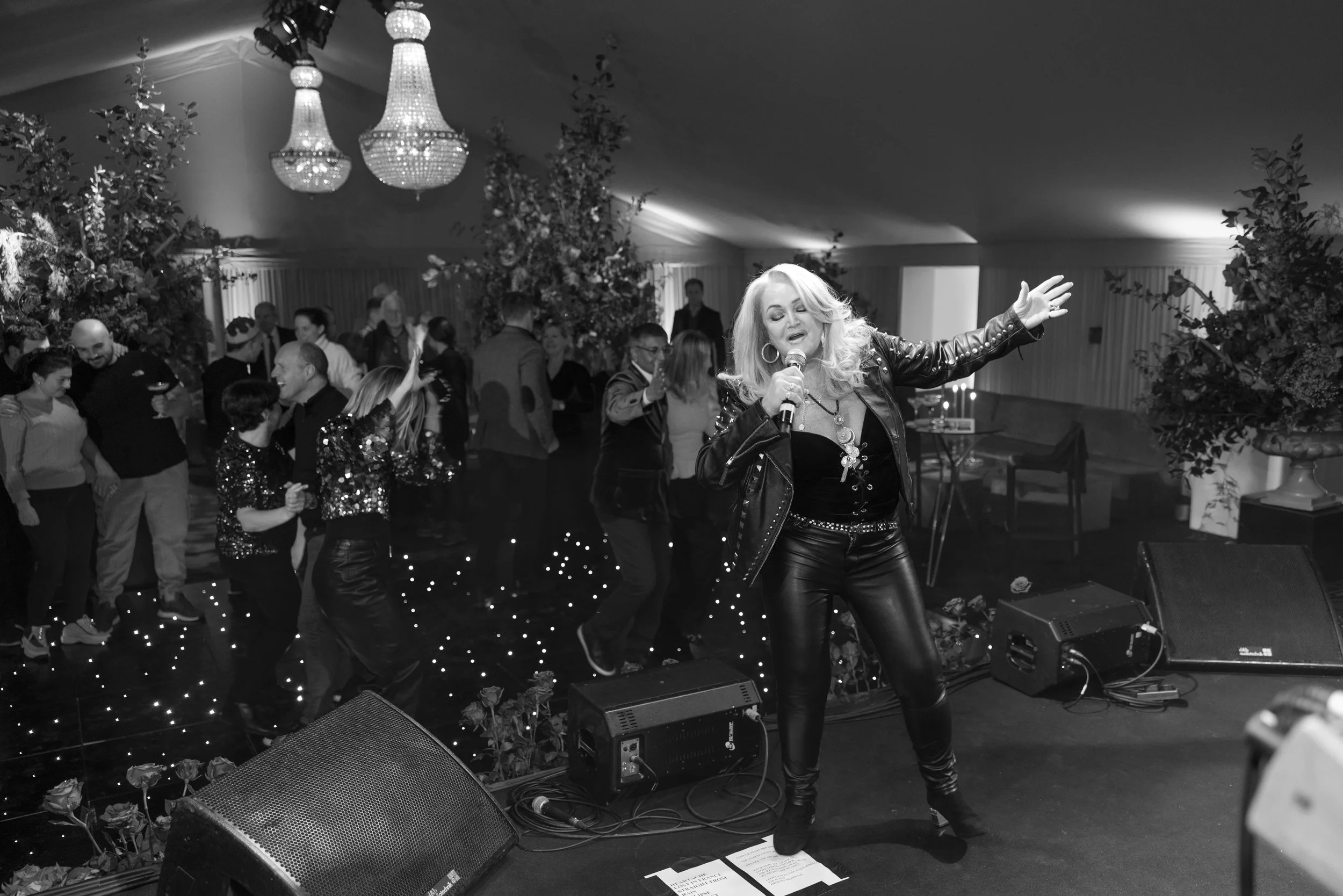 Female singer performing on stage at a party or event, with people dancing in the background.