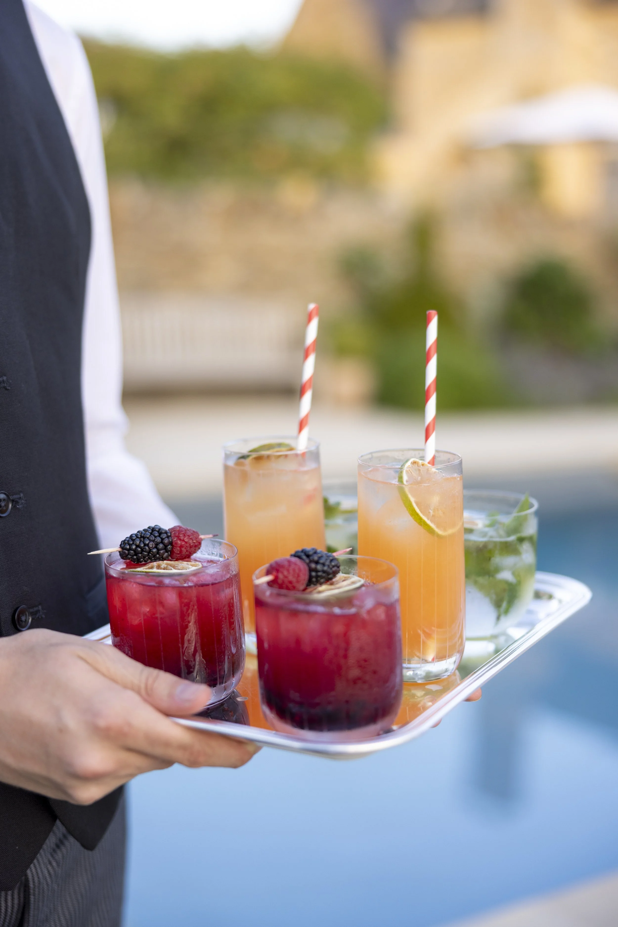 A server holding a tray with four colorful cocktails topped with fresh berries and lime slices, some of which have striped straws, set against a blurred outdoor background.