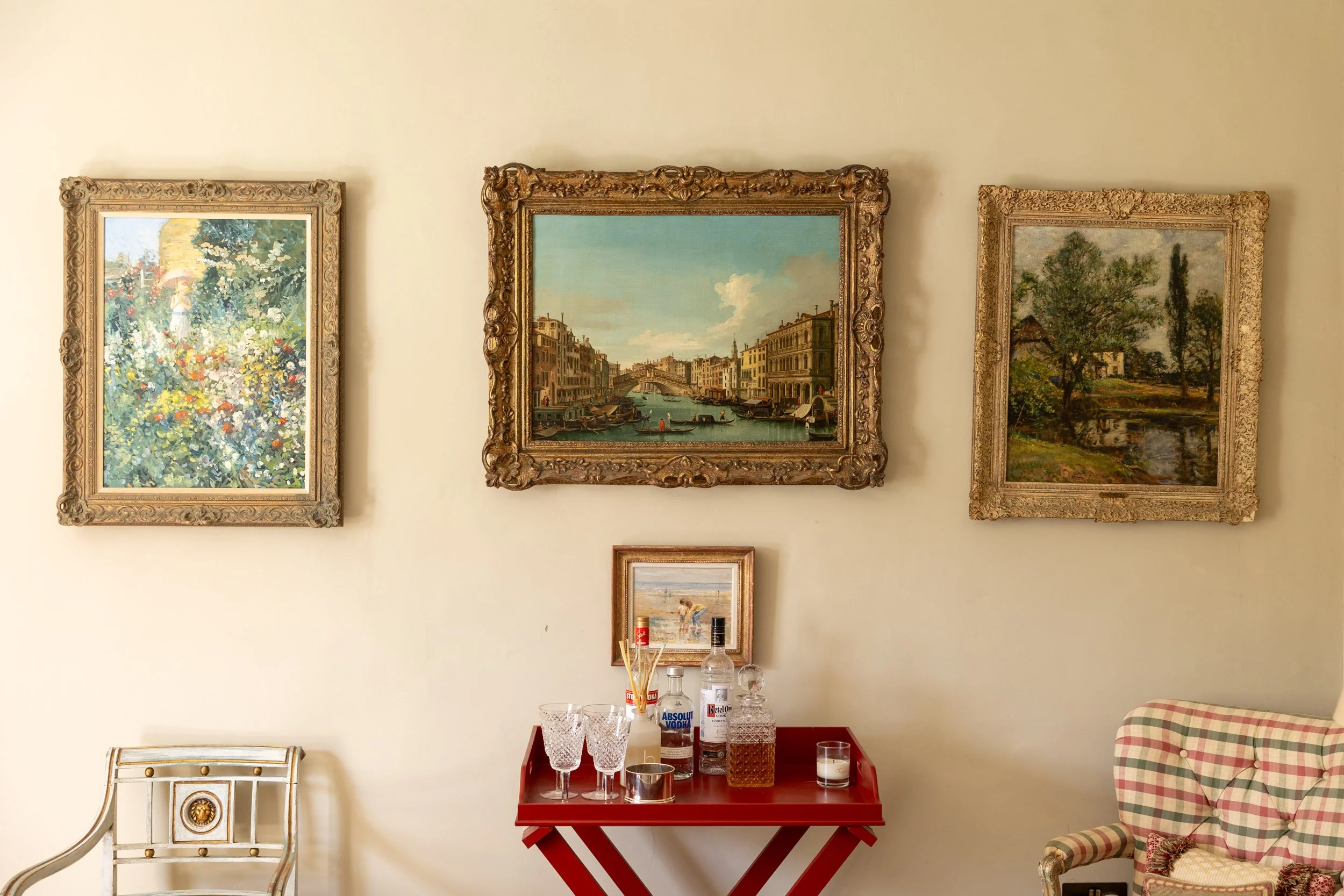 A wall with three framed paintings. The left painting depicts a garden scene, the middle shows a canal scene in Venice, Italy, and the right features a landscape with trees. Below the paintings, on the wall, there is a small table with bottles of alc