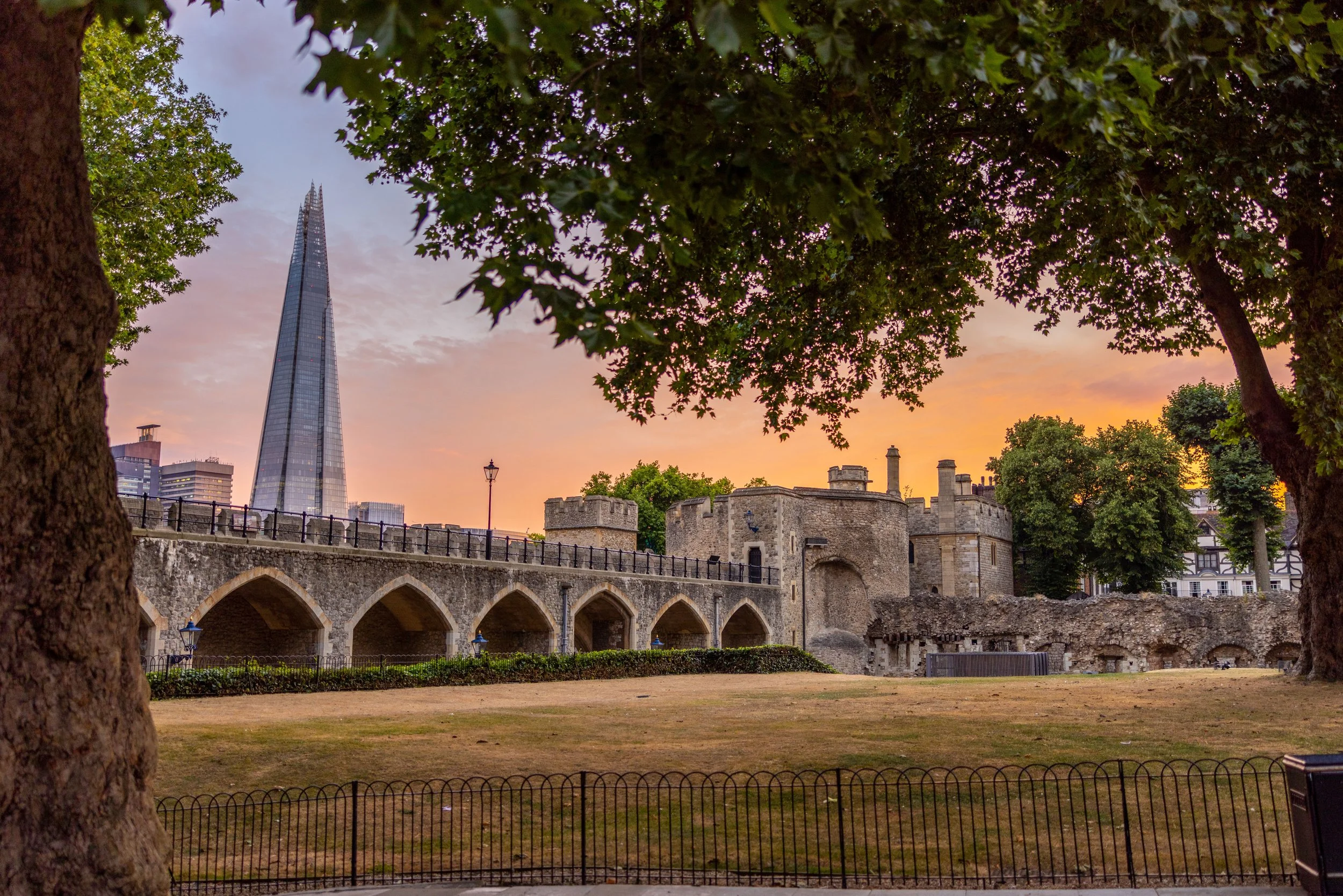 Sunset over a historic castle and modern skyscraper in London, with trees and a grassy area in the foreground.